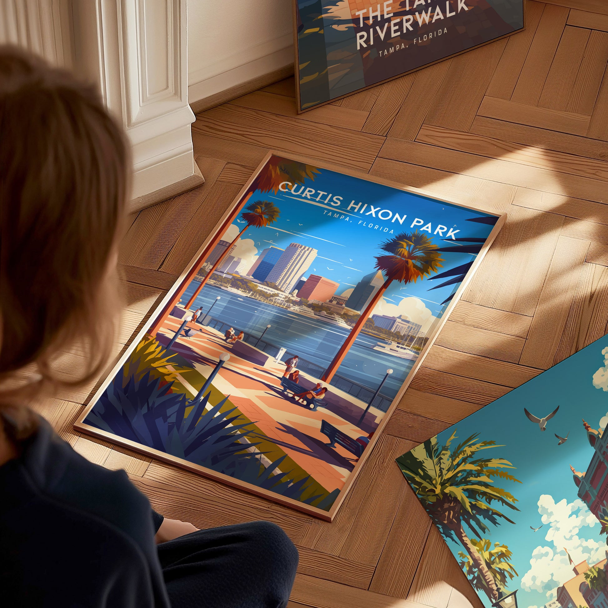 A person sitting on a wooden floor looks at a vibrant Curtis Hixon Park poster, depicting a sunny Tampa waterfront scene with palm trees and people enjoying the park.