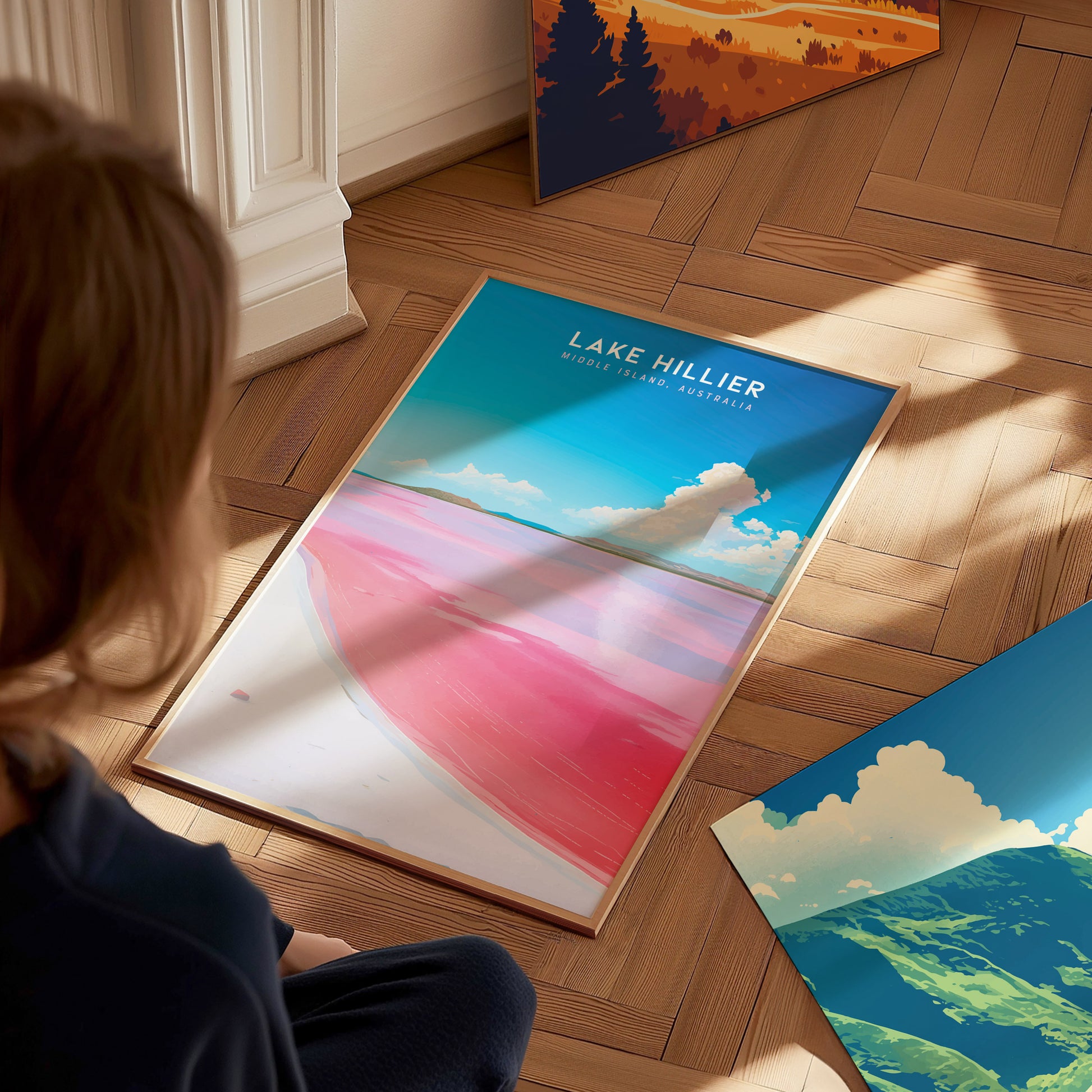 A person sits on a wooden floor, looking at a framed Lake Hillier poster featuring vibrant pink water and a blue sky. Other colorful posters are partially visible nearby.