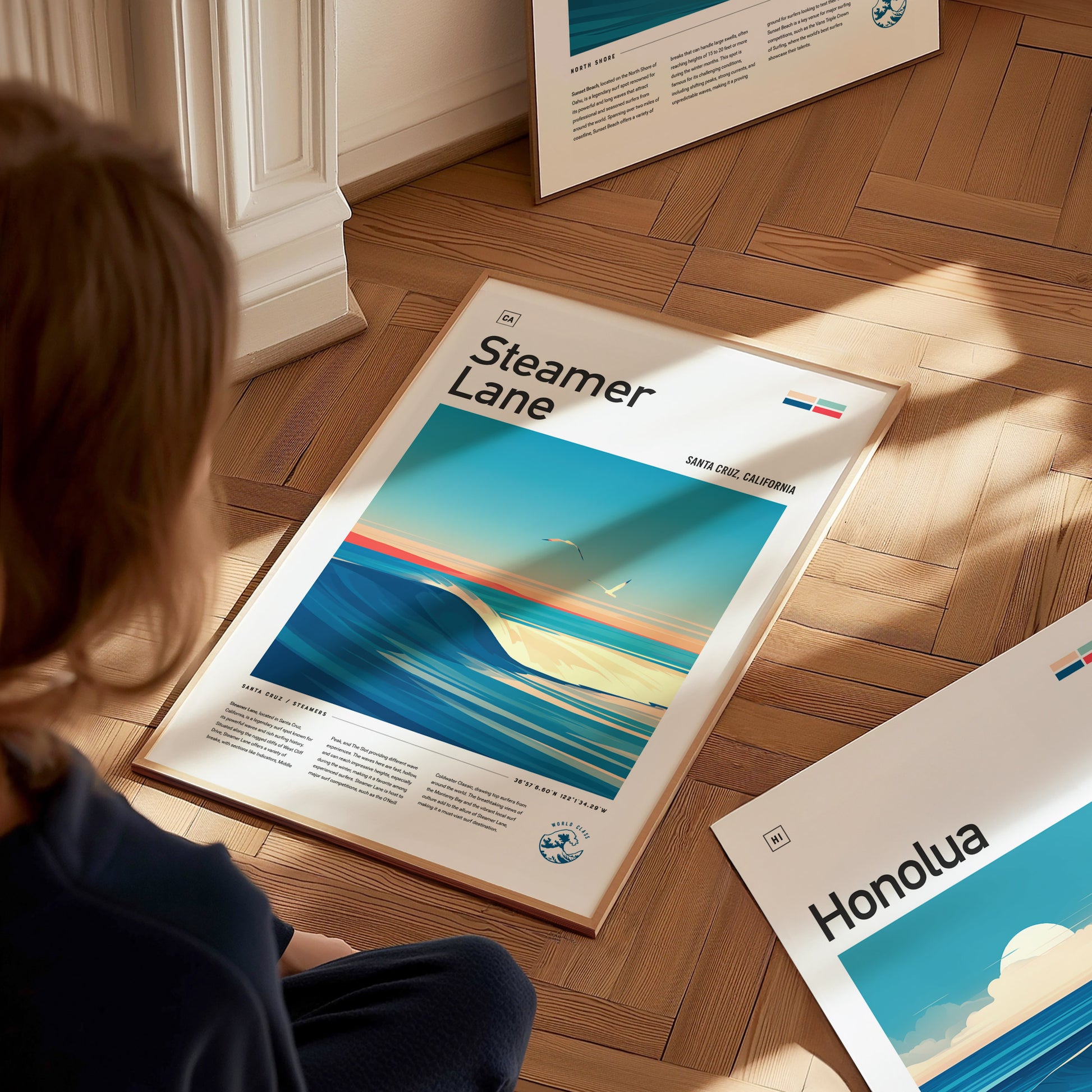A person is sitting on a wooden floor, looking at a framed poster titled Steamer Lane featuring an ocean wave scene. Another poster titled Honolua is partially visible nearby.