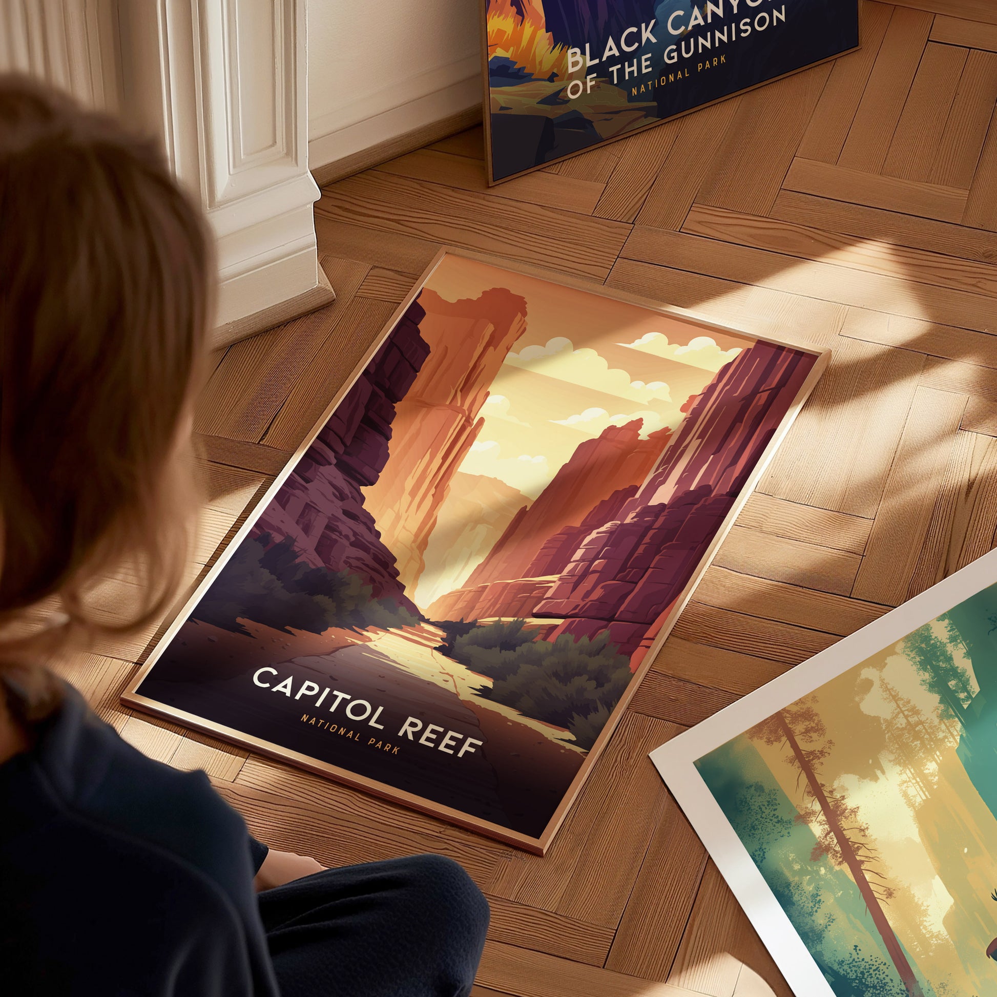 A person sits on a wooden floor, looking at a Capitol Reef National Park poster depicting a colorful canyon scene. Other posters, including one for Black Canyon, are nearby.