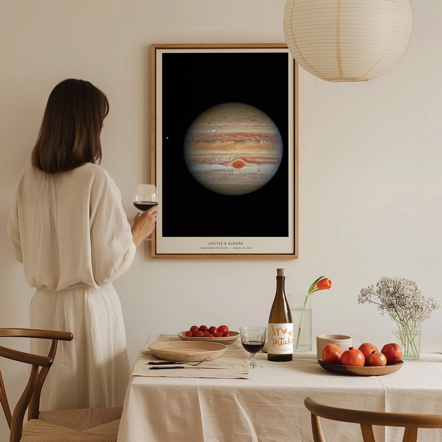 Woman in a white dress standing in a dining room with a framed poster of Jupiter on the wall.
