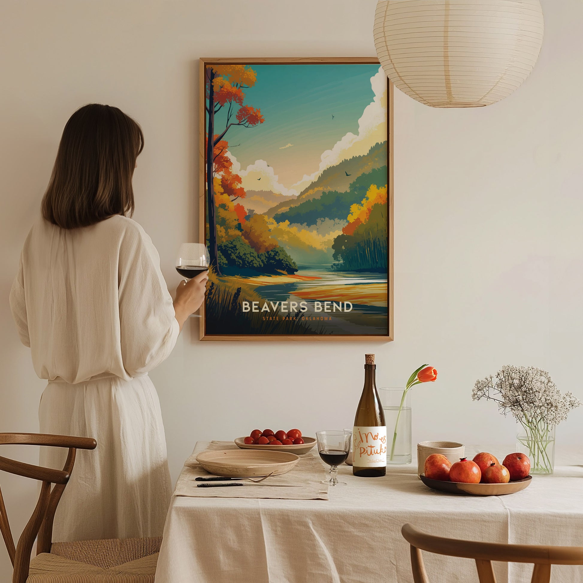 A woman in a white dress holds a glass of wine, admiring a framed Beavers Bend State Park poster. A dining table with fruit, wine, and flowers is in the.