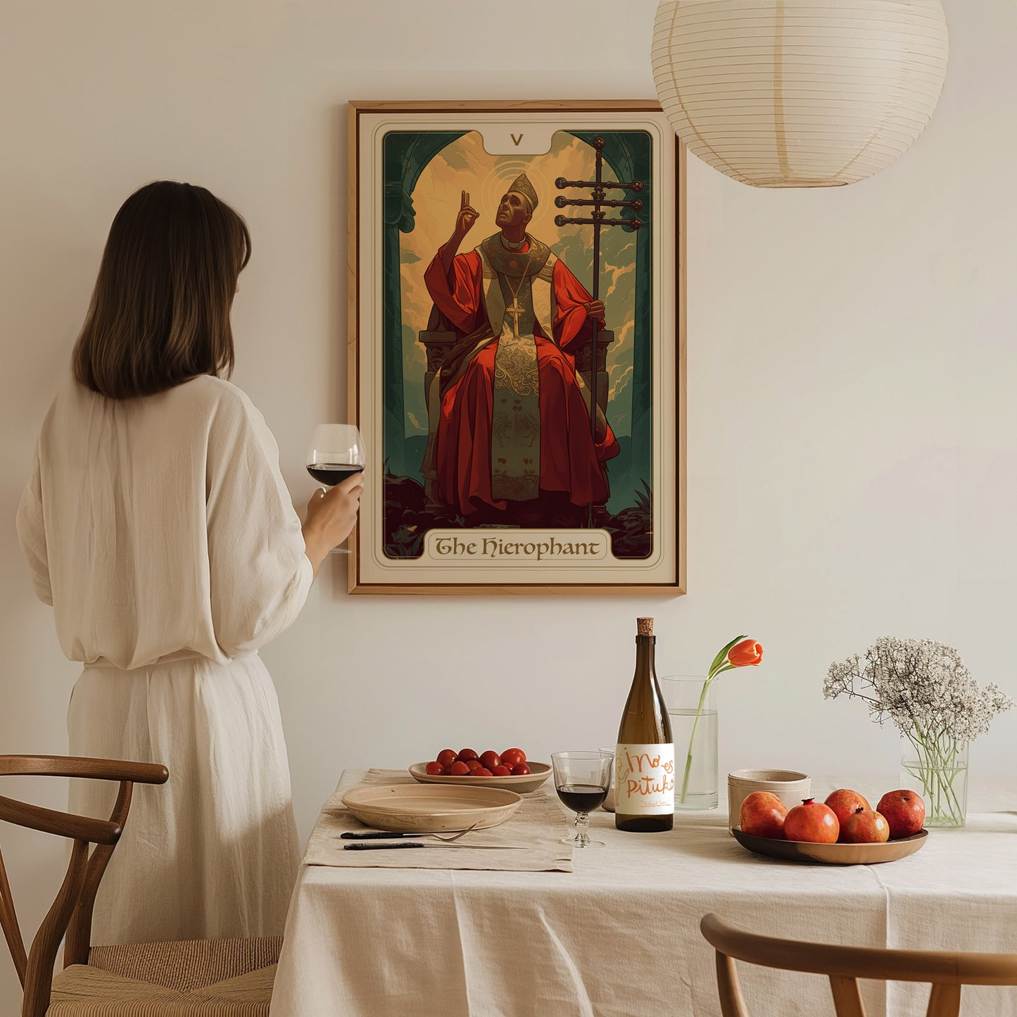 A woman in a white dress holds a glass of wine, admiring a Hierophant tarot poster on the wall. A table with fruit, wine, and flowers is in the foreground.