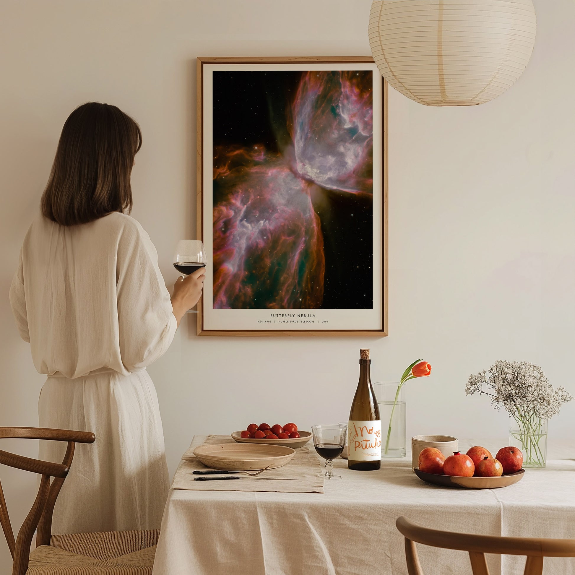 A woman in a white dress stands at a dining table, holding a glass of wine, while admiring a colorful Butterfly Nebula poster framed on the wall.