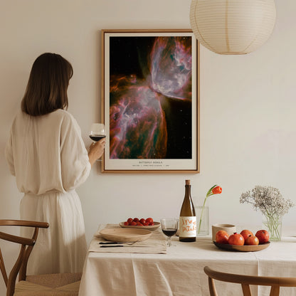 A woman in a white dress stands at a dining table, holding a glass of wine, while admiring a colorful Butterfly Nebula poster framed on the wall.