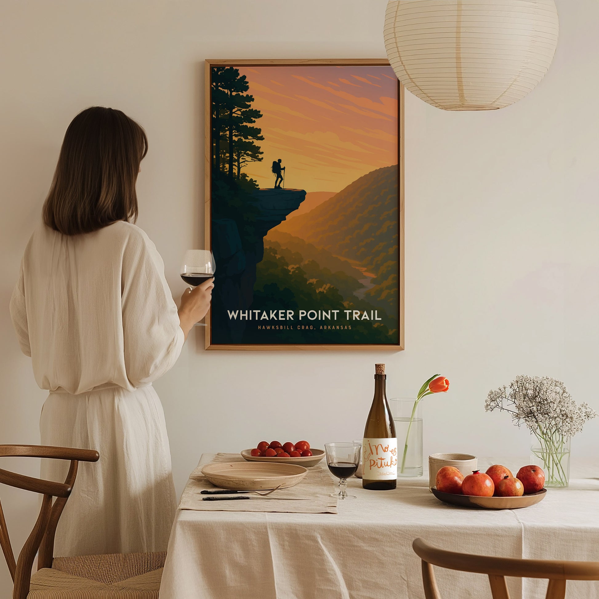 A woman in a white dress stands at a dining table, holding a glass of red wine, while admiring a framed art print of Whitaker Point Trail featuring a sunset over a mountainous landscape.