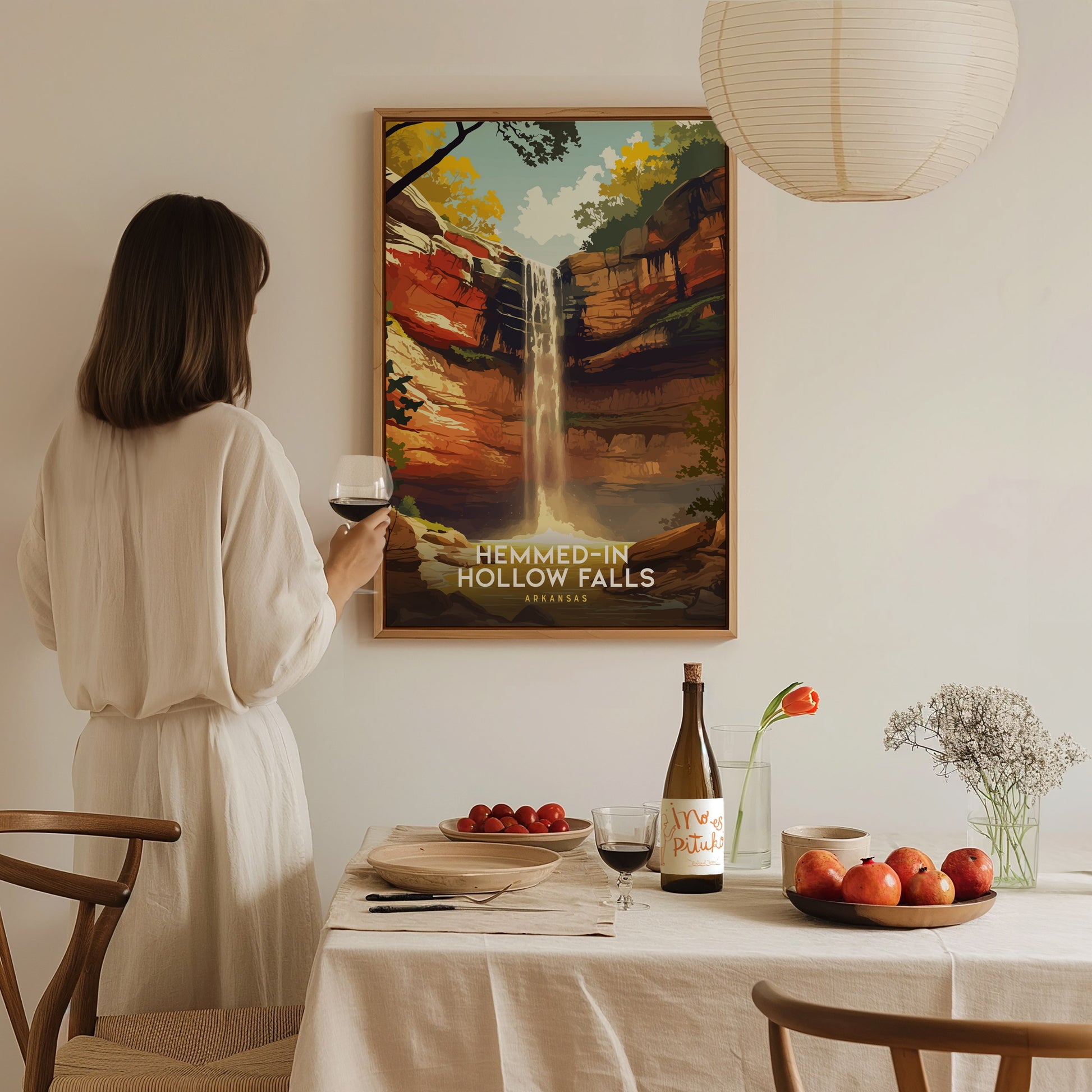 A woman in a light-colored dress stands at a dining table, holding a glass of wine. Behind her, a framed poster of Hemmed-In Hollow Falls showcases a waterfall surrounded by vibrant trees, creating a serene atmosphere in the room.