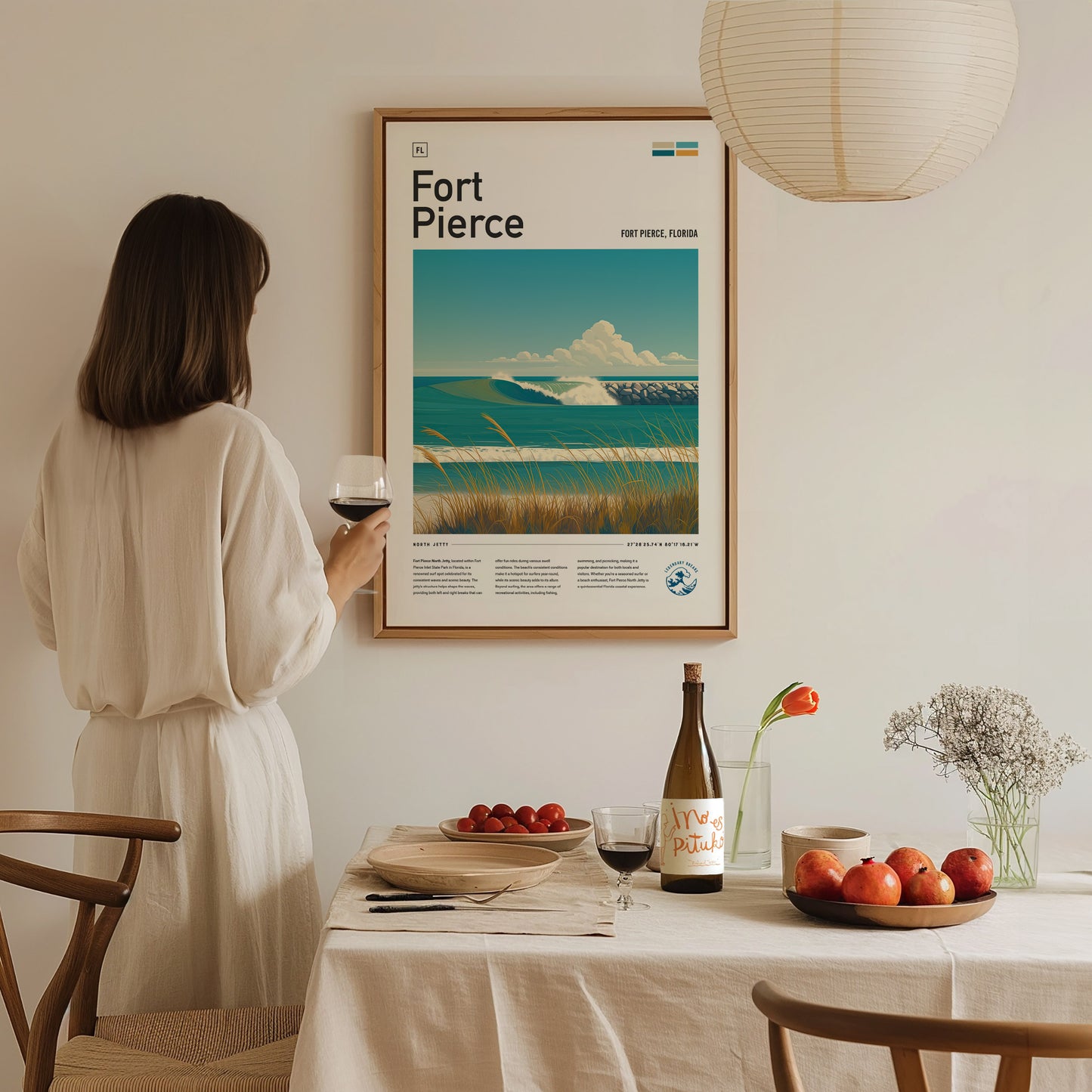 A woman in a white dress holds a wine glass, admiring a Fort Pierce surf spot poster on the wall. A table with fruit and wine is set below.