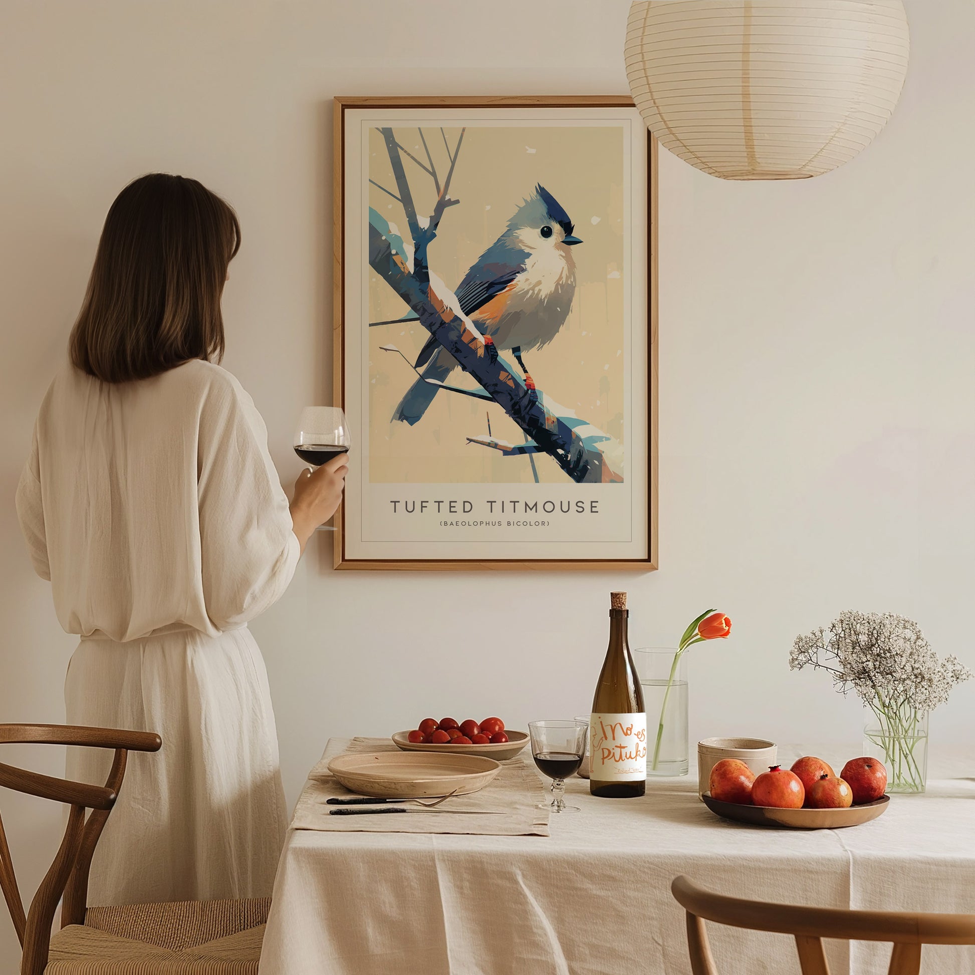 A woman in a white dress stands holding a glass of wine, admiring a framed Tufted Titmouse poster on the wall. A table set with fruit, wine, and flowers creates a cozy, elegant dining atmosphere.