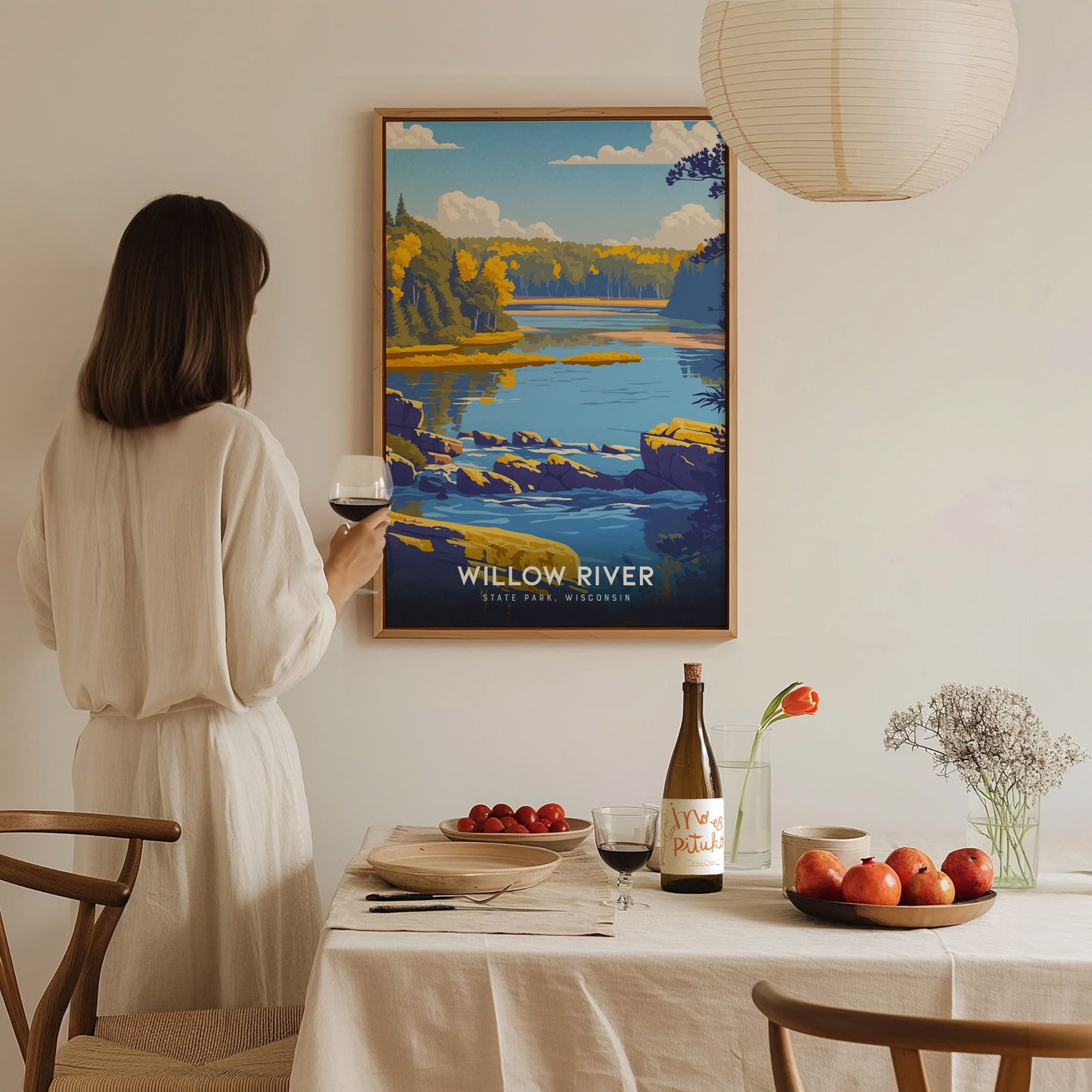 A woman in a light-colored, flowing outfit stands by a dining table, holding a glass of wine while gazing at a colorful framed poster of Willow River State Park.