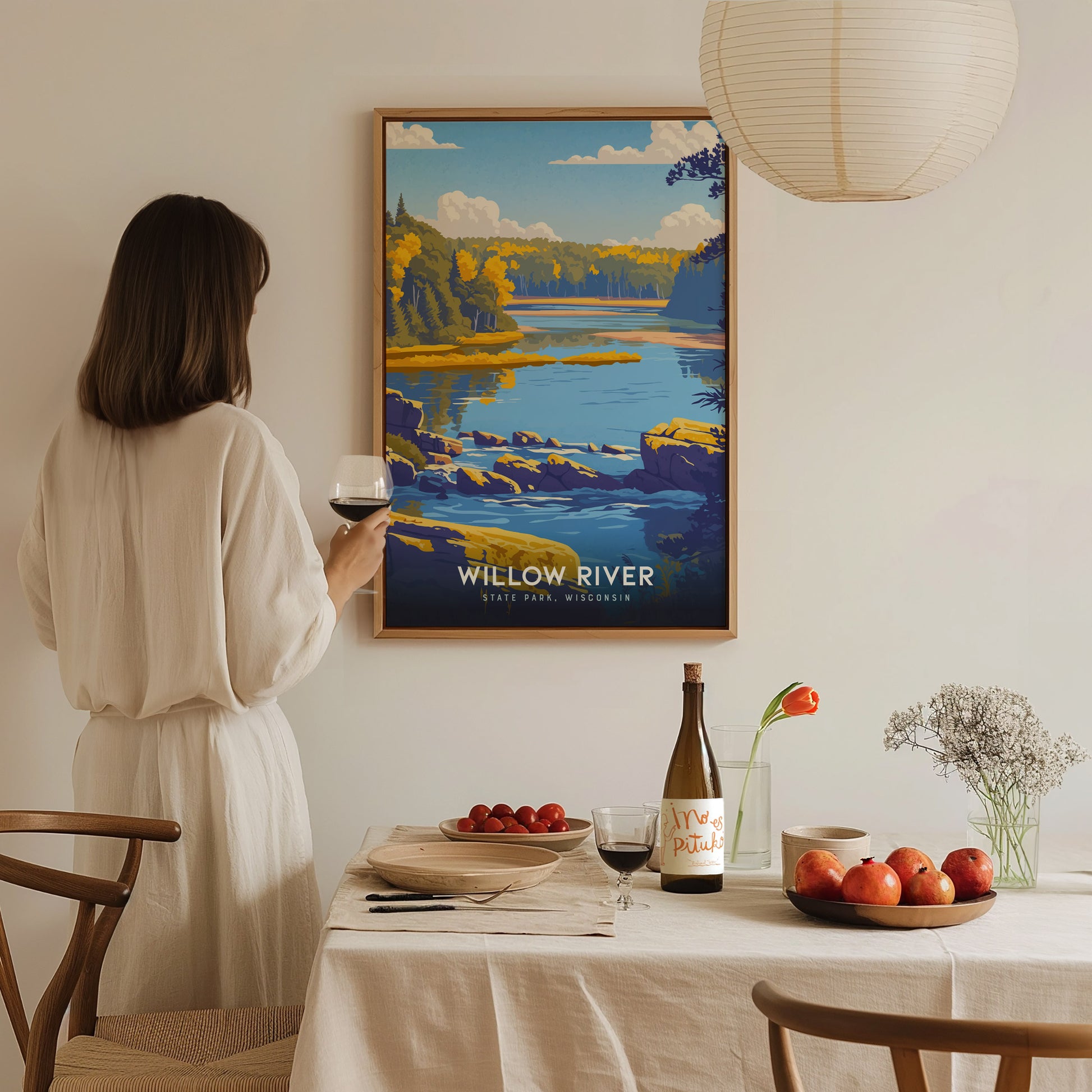 A woman in a light-colored, flowing outfit stands by a dining table, holding a glass of wine while gazing at a colorful framed poster of Willow River State Park.