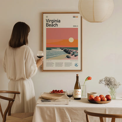 A woman in a white dress admires a framed Virginia Beach poster with a sunset design. A dining table with wine, fruit, and flowers is in the foreground.