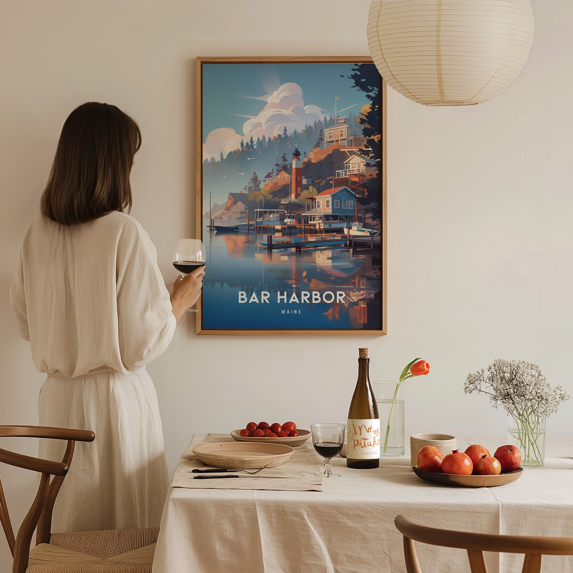 A woman in a white dress holds a glass of wine, admiring a Bar Harbor, Maine poster on the wall. A table with fruit and wine is in the foreground.