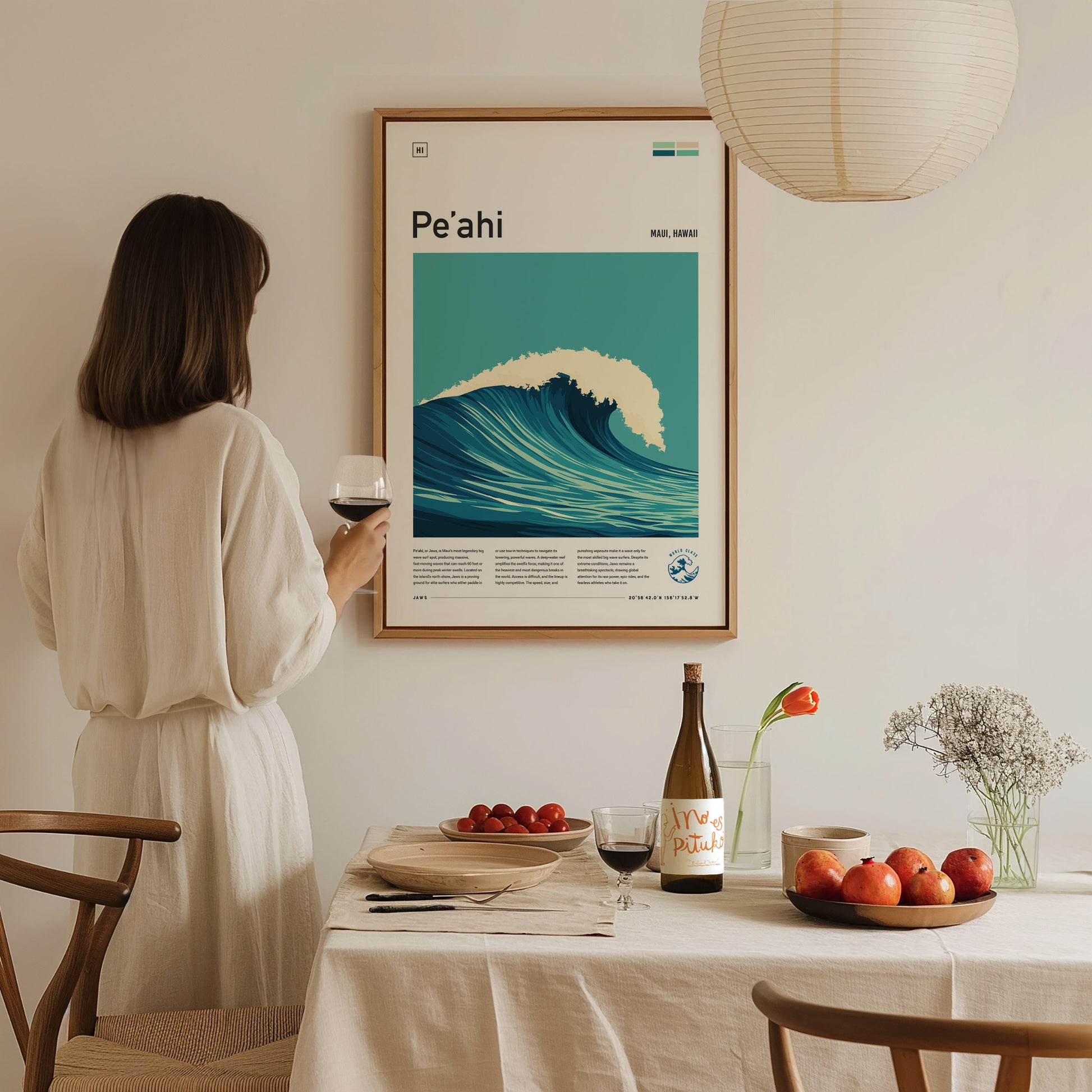 A woman in a white dress admires a framed Peʻahi surf poster on a wall above a dining table with wine, apples, and flowers.