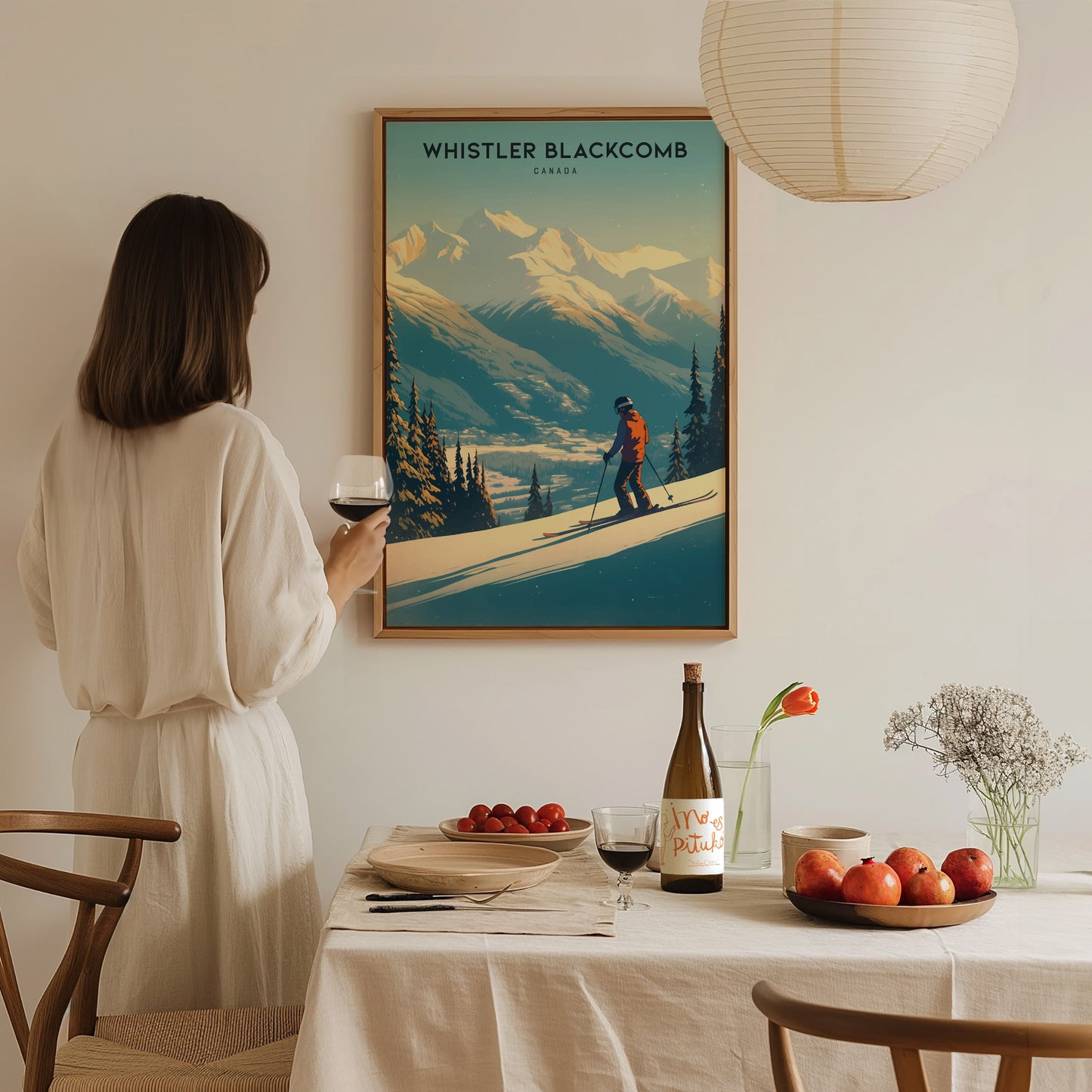 A woman in a white dress holds a glass of wine, admiring a Whistler Blackcomb ski resort poster. The dining table features fruit, wine, and a small floral arrangement.