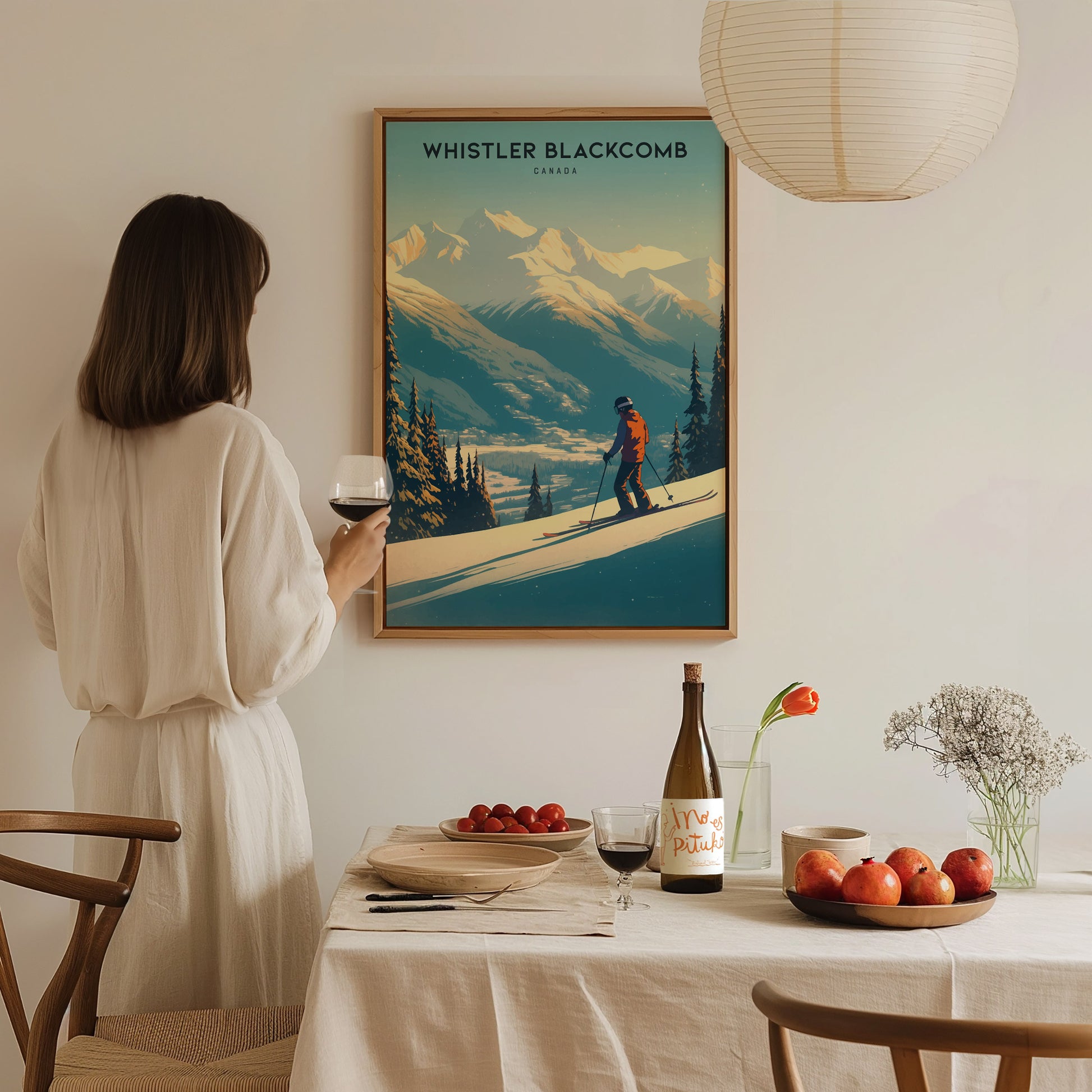 A woman in a white dress holds a glass of wine, admiring a Whistler Blackcomb ski resort poster. The dining table features fruit, wine, and a small floral arrangement.