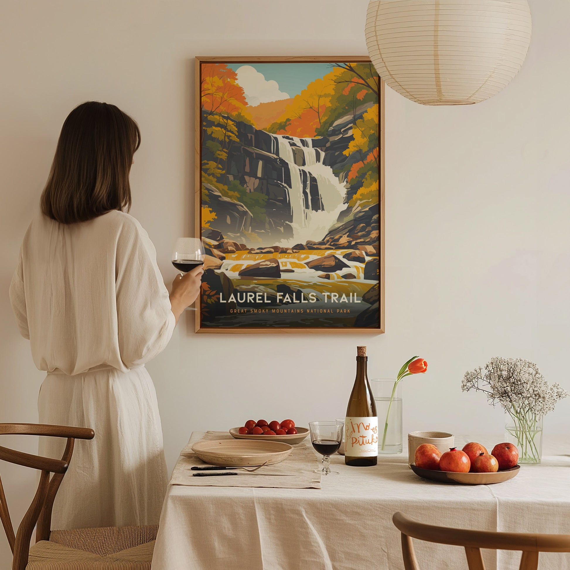 A woman in a white dress holds a glass of wine while admiring a Laurel Falls Trail poster. The table is set with fruit, wine, and a vase with flowers.