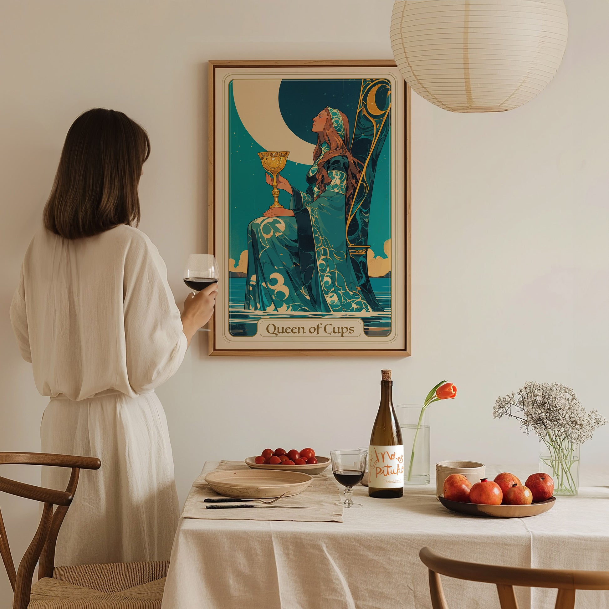 A woman in a light-colored dress stands at a dining table, holding a glass of wine, while gazing at a colorful tarot card poster titled Queen of Cups on the wall.