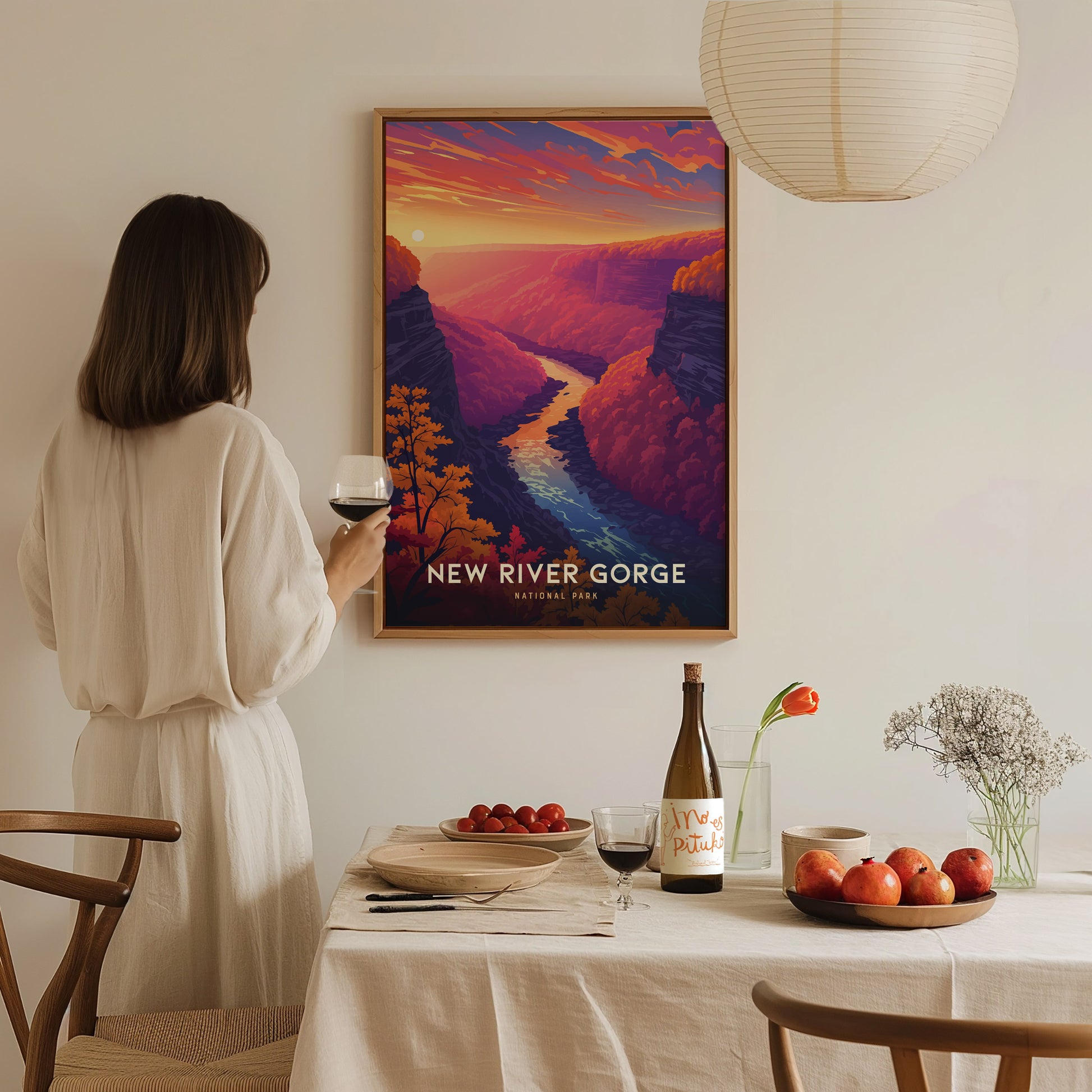 A woman in a white dress admires a colorful New River Gorge poster on a wall. A table with wine, apples, and flowers is in the foreground, creating a cozy.