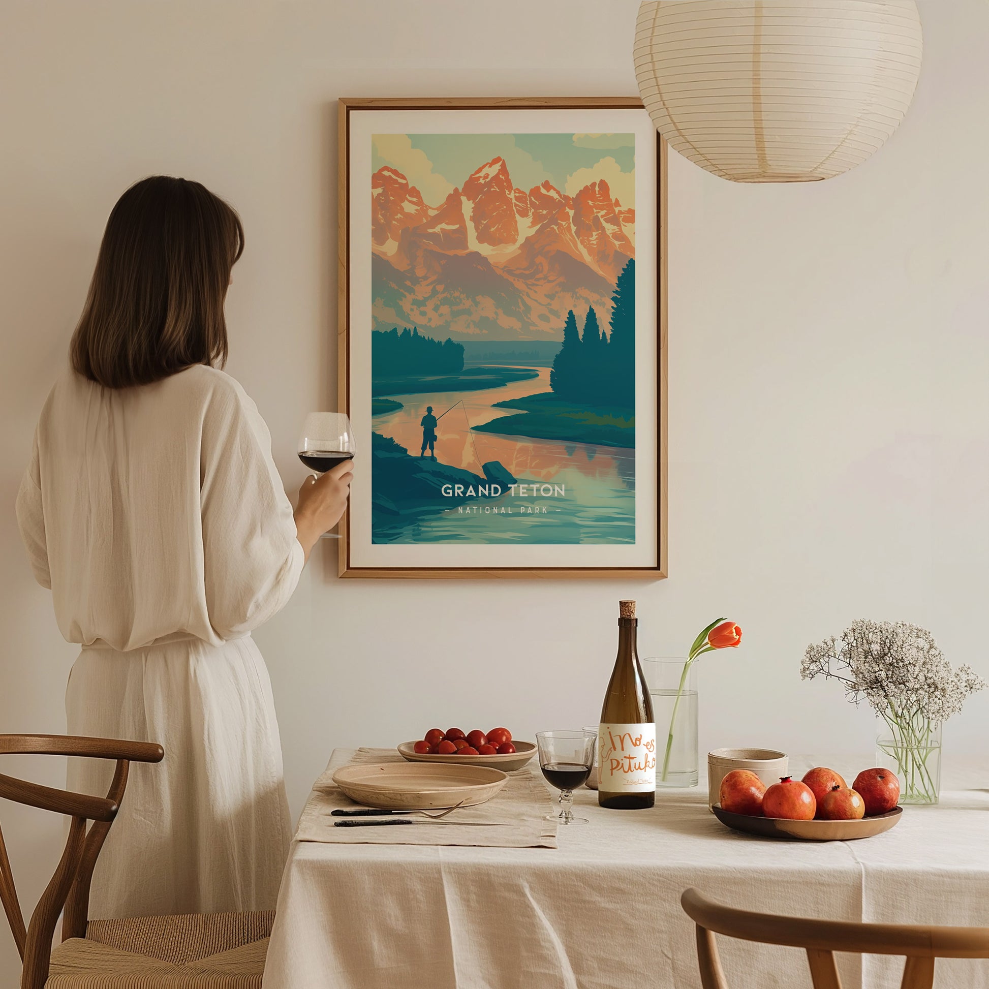 A woman in a white dress admires a framed Grand Teton National Park poster on the wall, with a dining table set with wine, fruit, and flowers in the foreground.