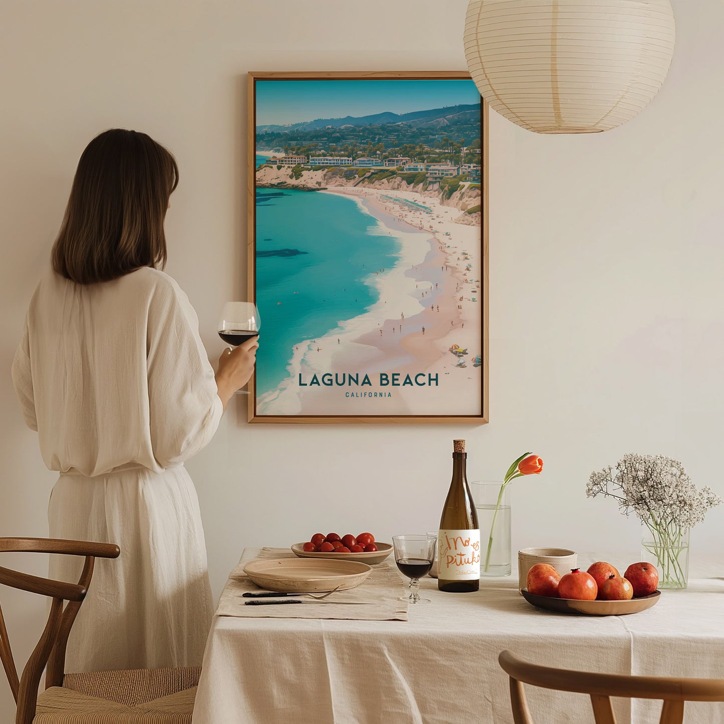 A woman in a white dress holds a glass of wine, admiring a framed Laguna Beach poster. A dining table is set with fruit, a bottle, and flowers.