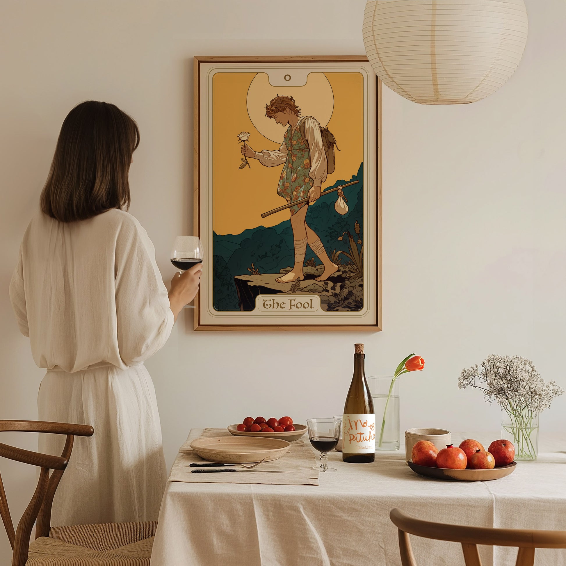 A woman in a white dress holds a glass of wine, gazing at a framed Fool tarot card poster on the wall. A table with fruit and wine sits nearby.