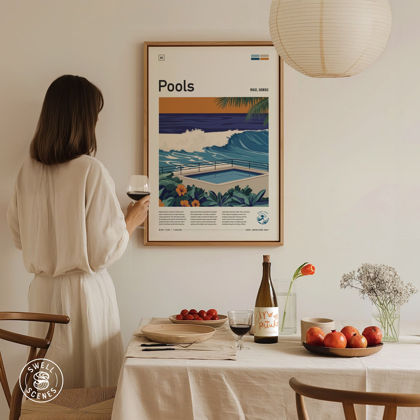 A woman in a white dress examines a framed surf poster featuring waves and a pool. A dining table below holds wine, apples, and flowers, creating a cozy atmosphere.