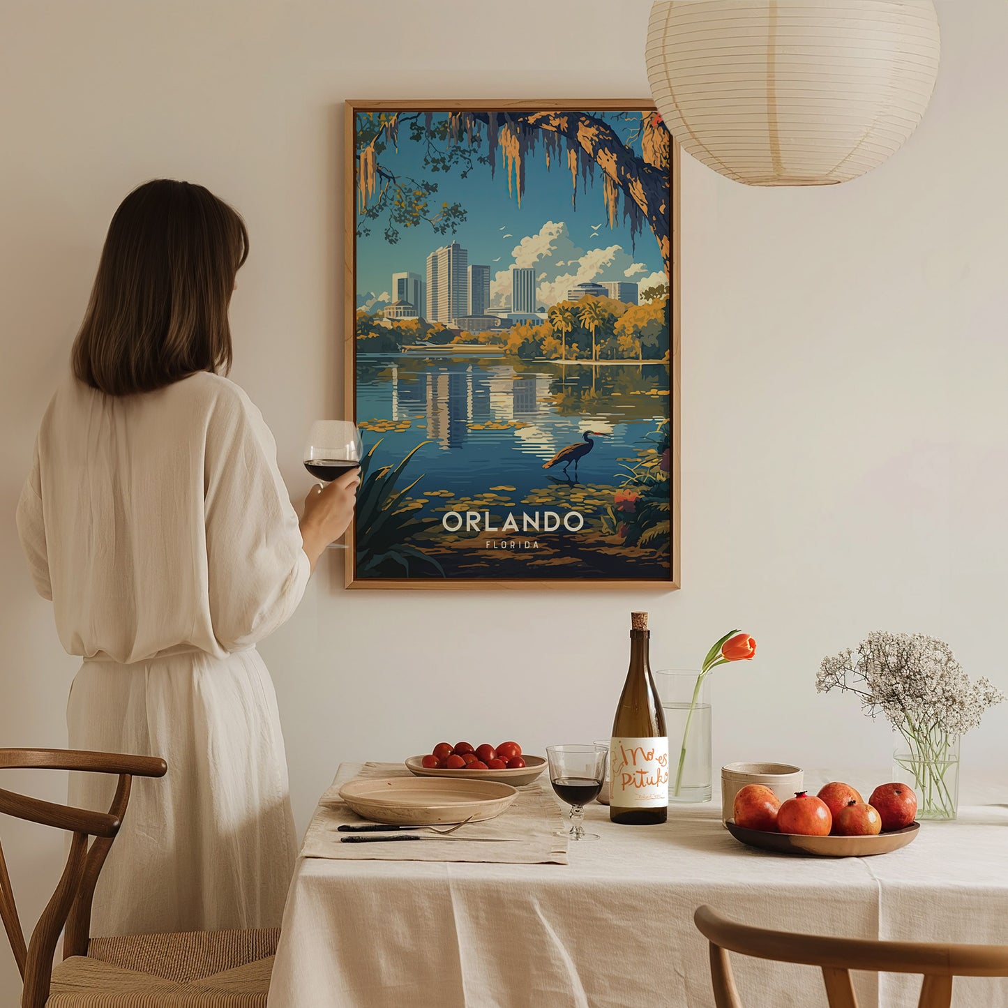 A woman in a white dress holds a glass of wine, admiring a framed Orlando skyline poster on a wall. A table with fruits, flowers, and wine is in the.