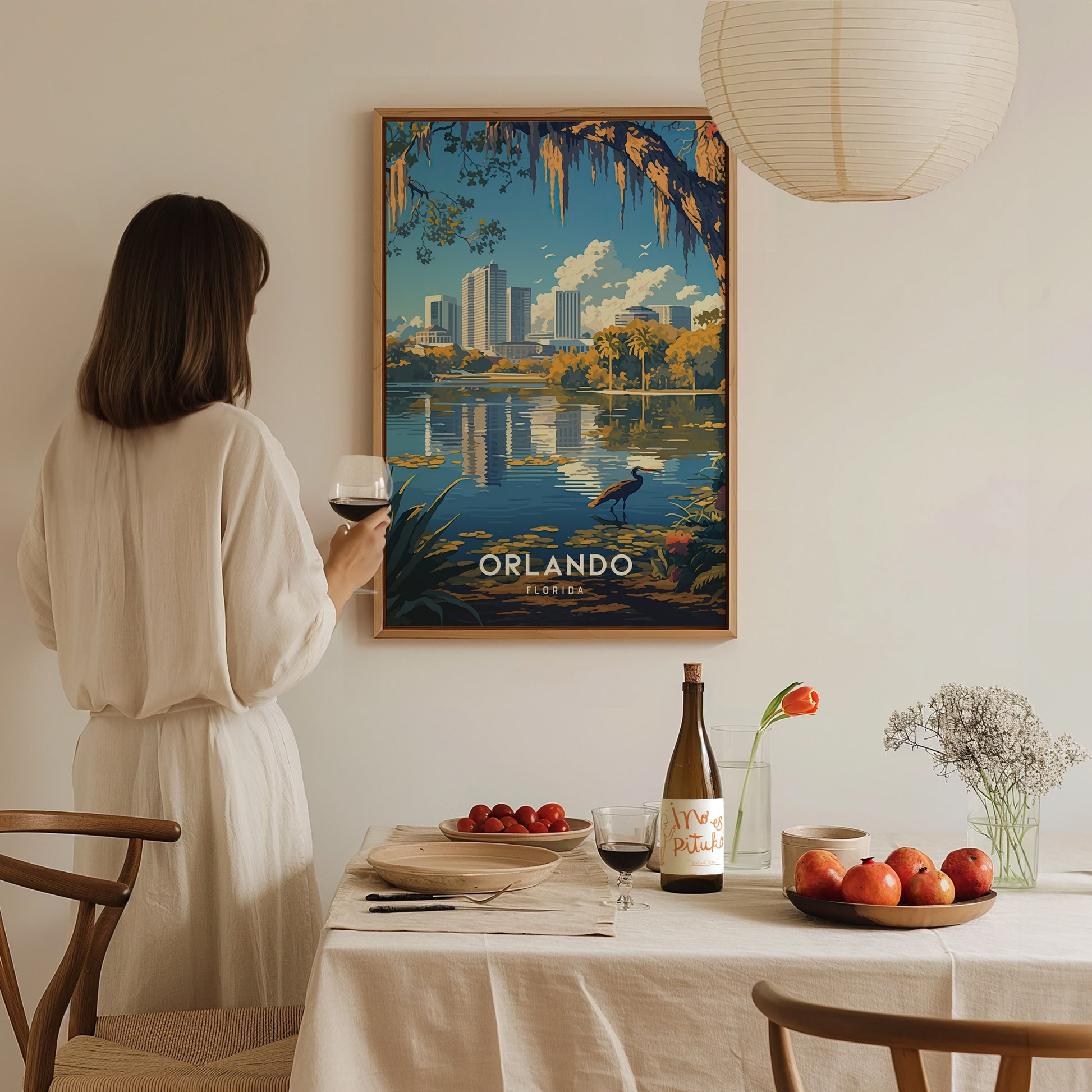 A woman in a white dress holds a glass of wine, admiring a framed Orlando skyline poster on a wall. A table with fruits, flowers, and wine is in the.