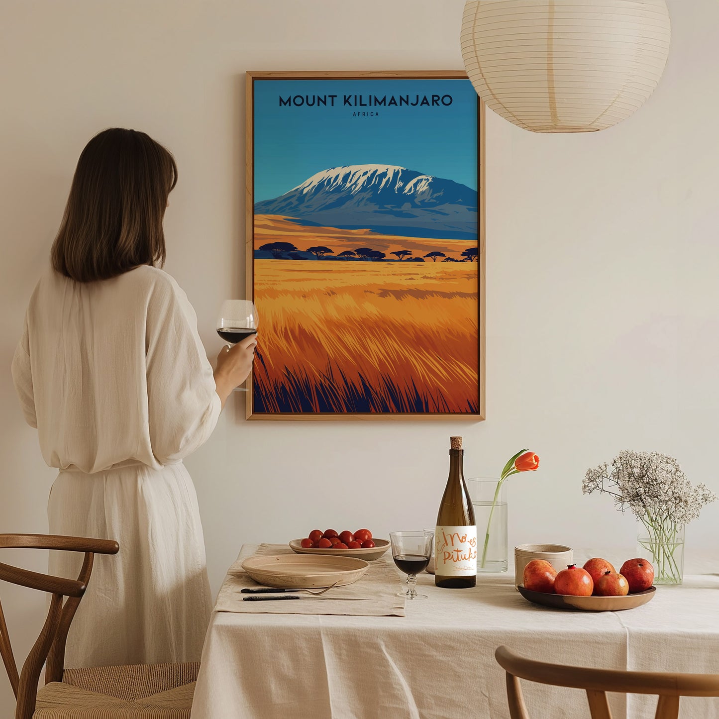 A woman in a white dress holds a wine glass, admiring a Mount Kilimanjaro poster on the wall. The dining table features fruits, wine,.