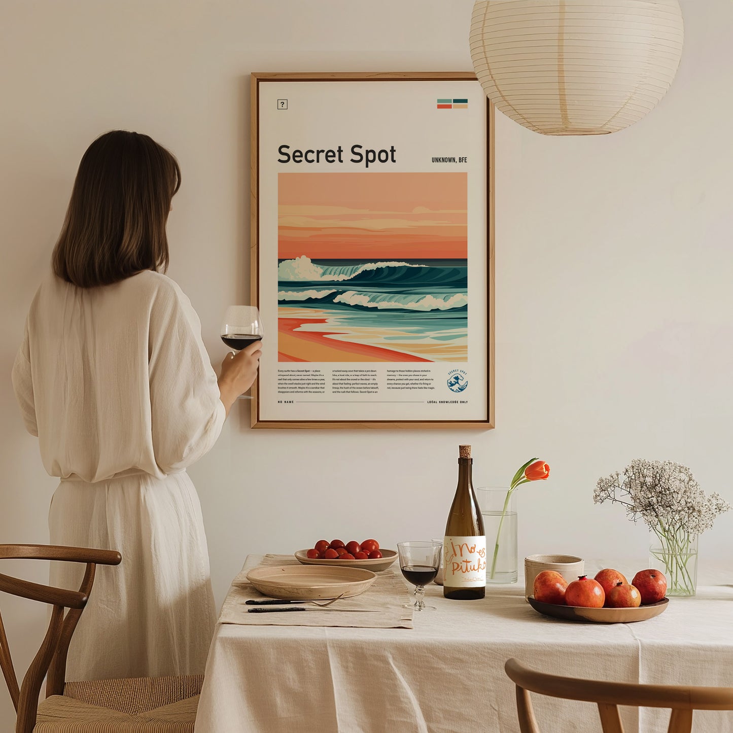 A person in a white dress admires a surf poster titled Secret Spot on the wall. The table below holds a wine bottle, glasses, and a bowl of fruit.
