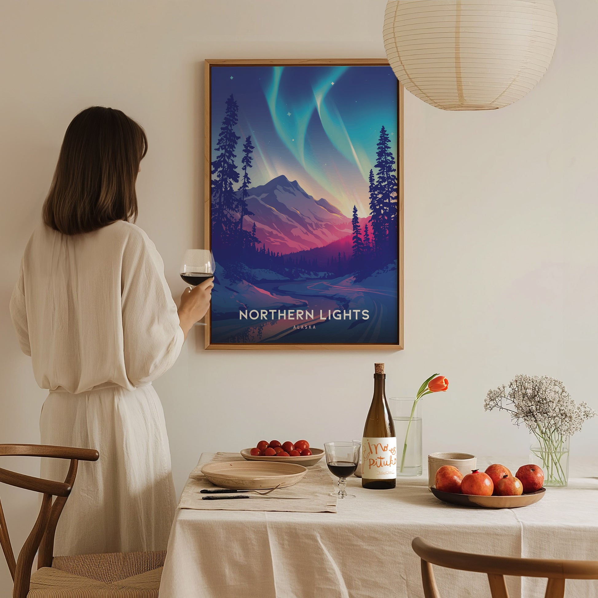 A woman in a white dress holds a glass of wine, admiring a framed Northern Lights Alaska poster on the wall. A table with a bottle, fruit, and flowers is.
