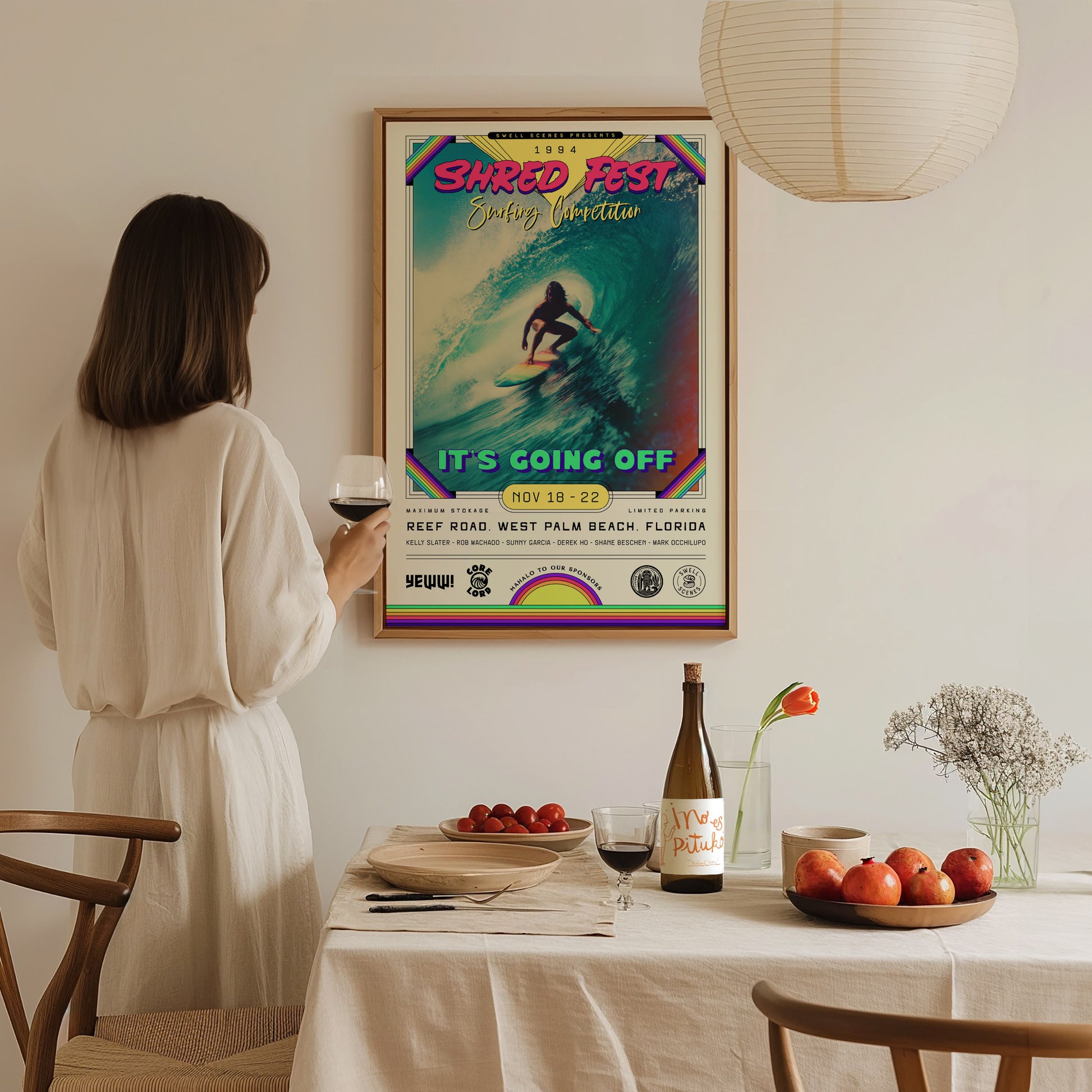 A woman stands in a cozy, well-lit dining space, holding a glass of wine and gazing at a colorful framed poster titled SHRED FEST, featuring a surfer riding a wave.