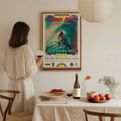 A woman stands in a cozy, well-lit dining space, holding a glass of wine and gazing at a colorful framed poster titled SHRED FEST, featuring a surfer riding a wave.