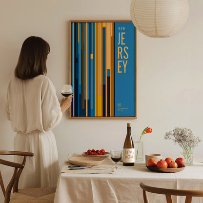 A woman in a white dress admires a colorful New Jersey poster on the wall, holding a glass of wine near a table set with fruit and flowers.