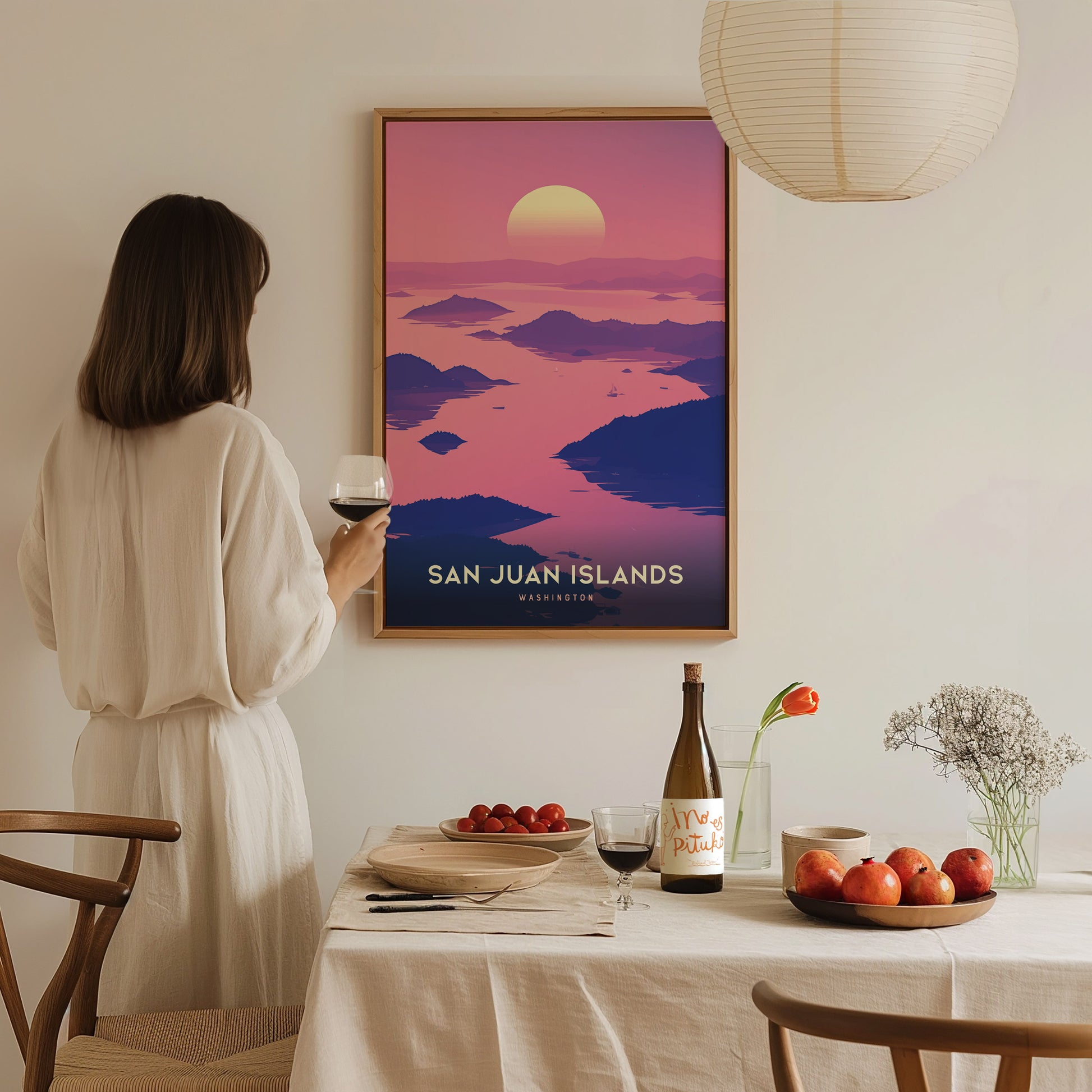 A woman in a white dress admires a San Juan Islands poster on the wall. A dining table with wine, fruit, and flowers is set in the foreground.