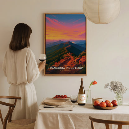 A woman in a white dress holds a glass of wine while admiring a colorful art print titled Franconia Ridge Loop, featuring a vibrant sunset over a mountainous landscape.