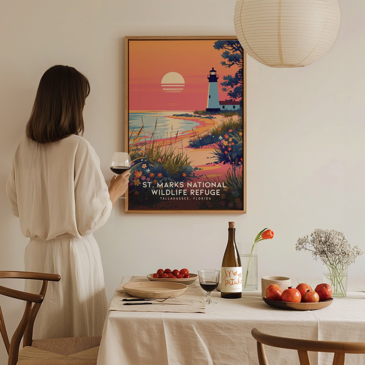 A person in a white dress admires a St. Marks National Wildlife Refuge poster depicting a lighthouse at sunset. A table with wine, fruit, and flowers is in the foreground.