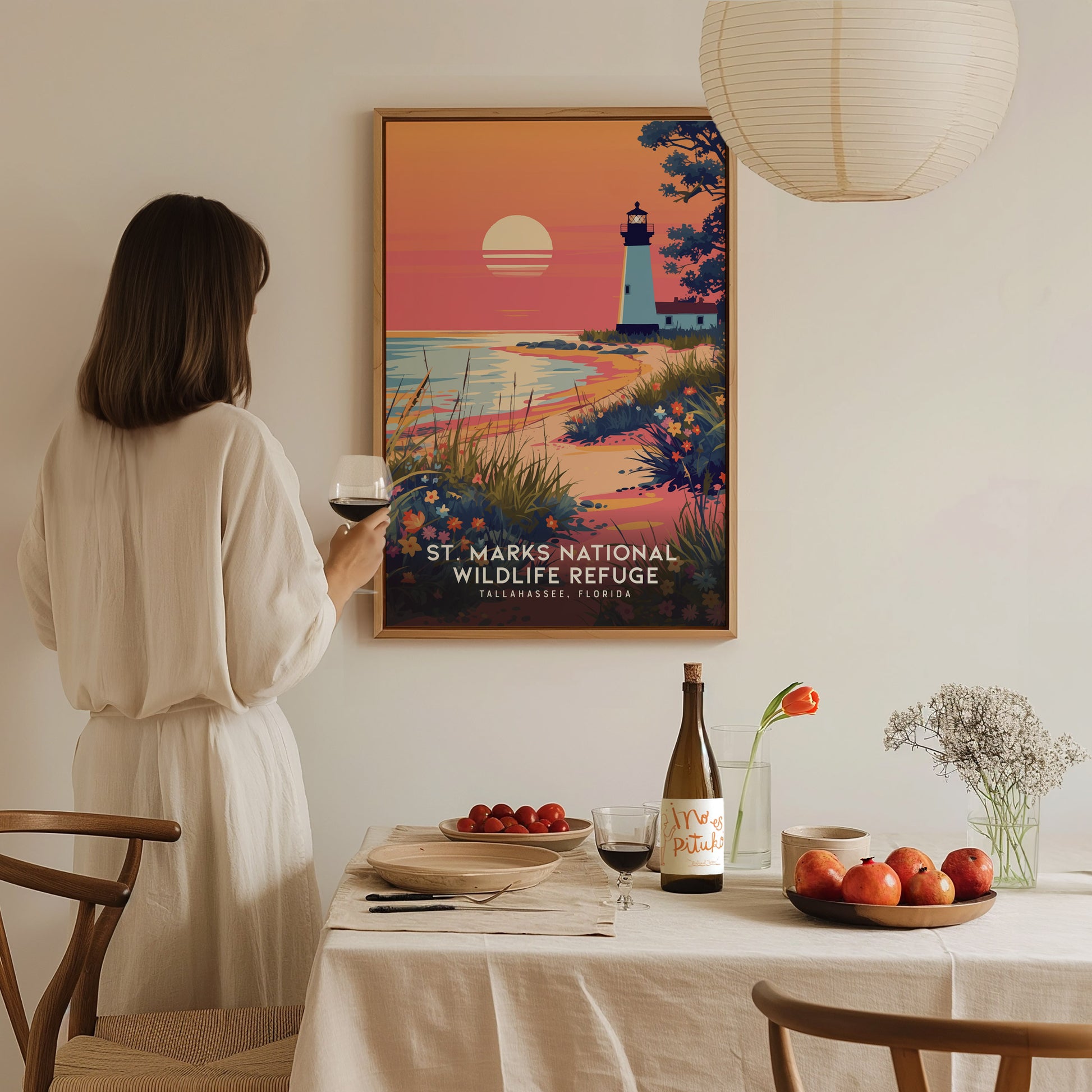 A person in a white dress admires a St. Marks National Wildlife Refuge poster depicting a lighthouse at sunset. A table with wine, fruit, and flowers is in the foreground.