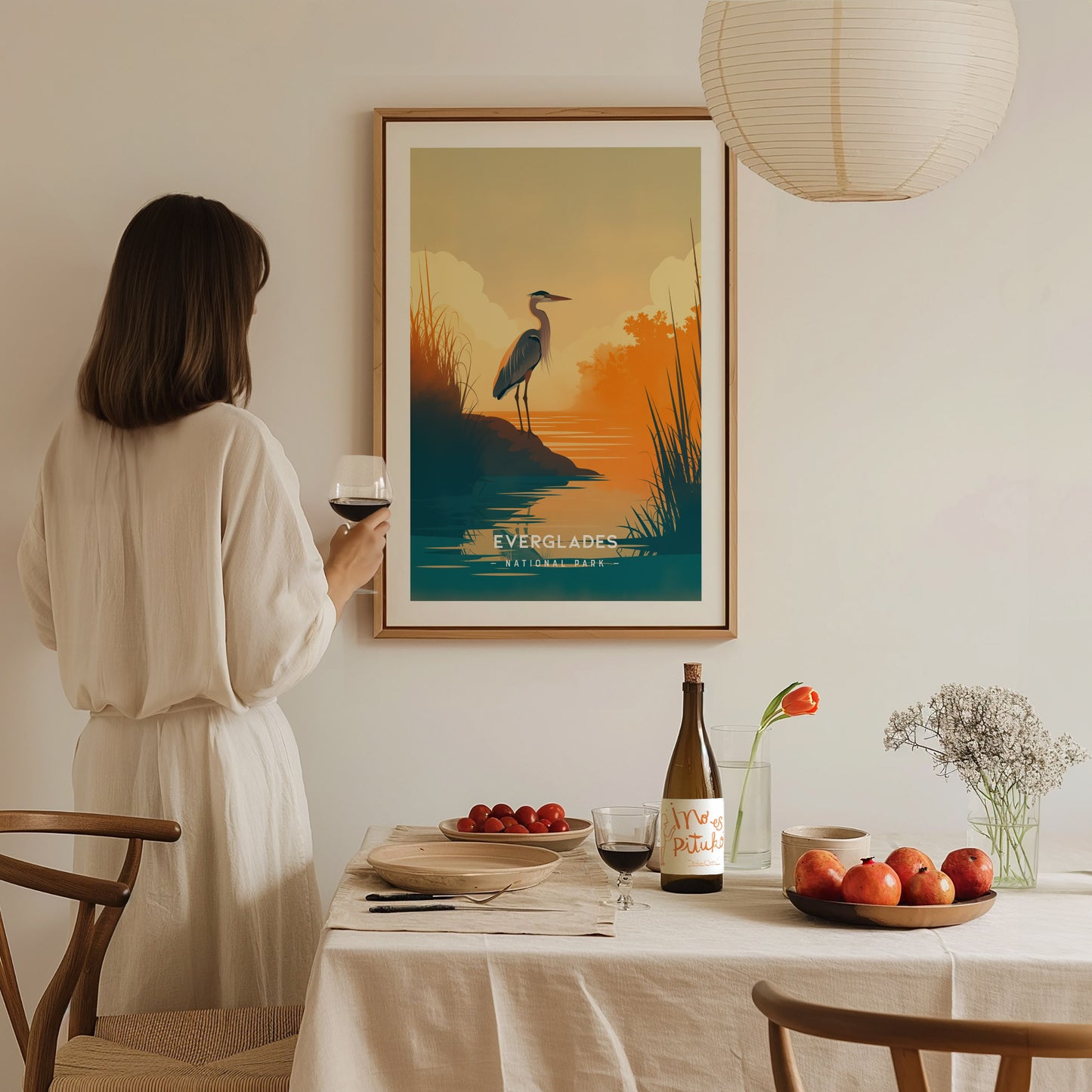 A woman in a white dress holds a glass of wine, admiring a framed Everglades poster on a wall. A table with fruit and a bottle is in the foreground.