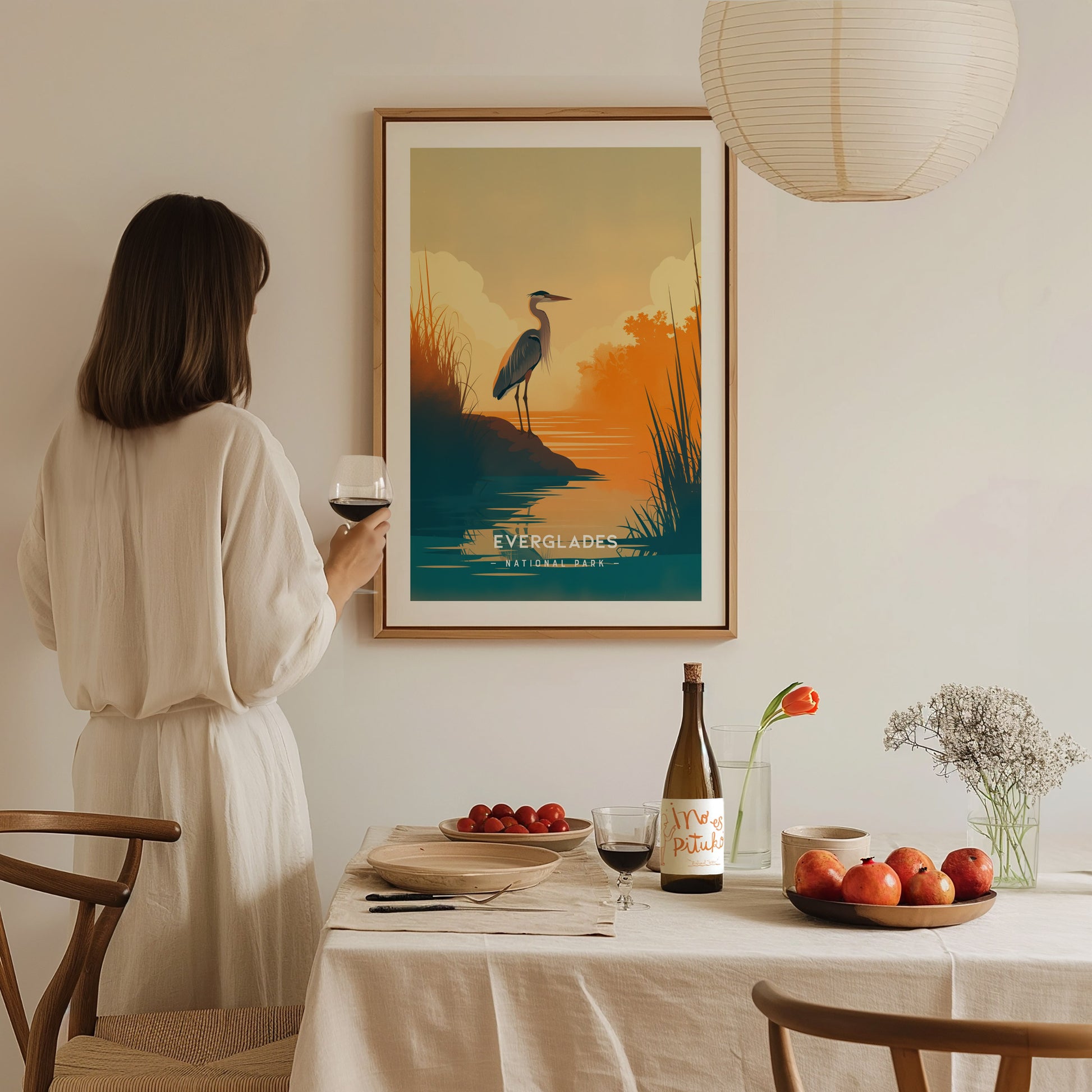 A woman in a white dress holds a glass of wine, admiring a framed Everglades poster on a wall. A table with fruit and a bottle is in the foreground.