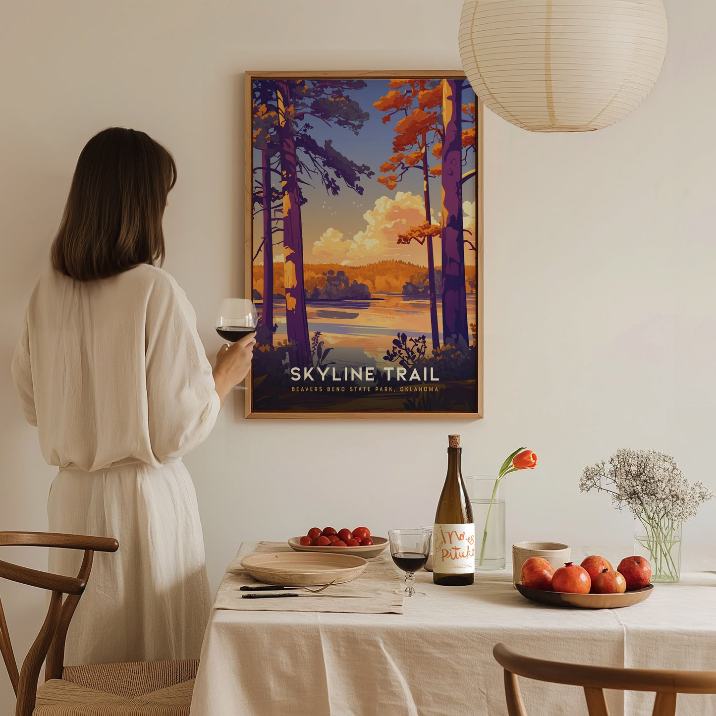 A woman in a white dress admires a colorful Skyline Trail poster on a wall. A table with wine, apples, and flowers is set in the foreground.