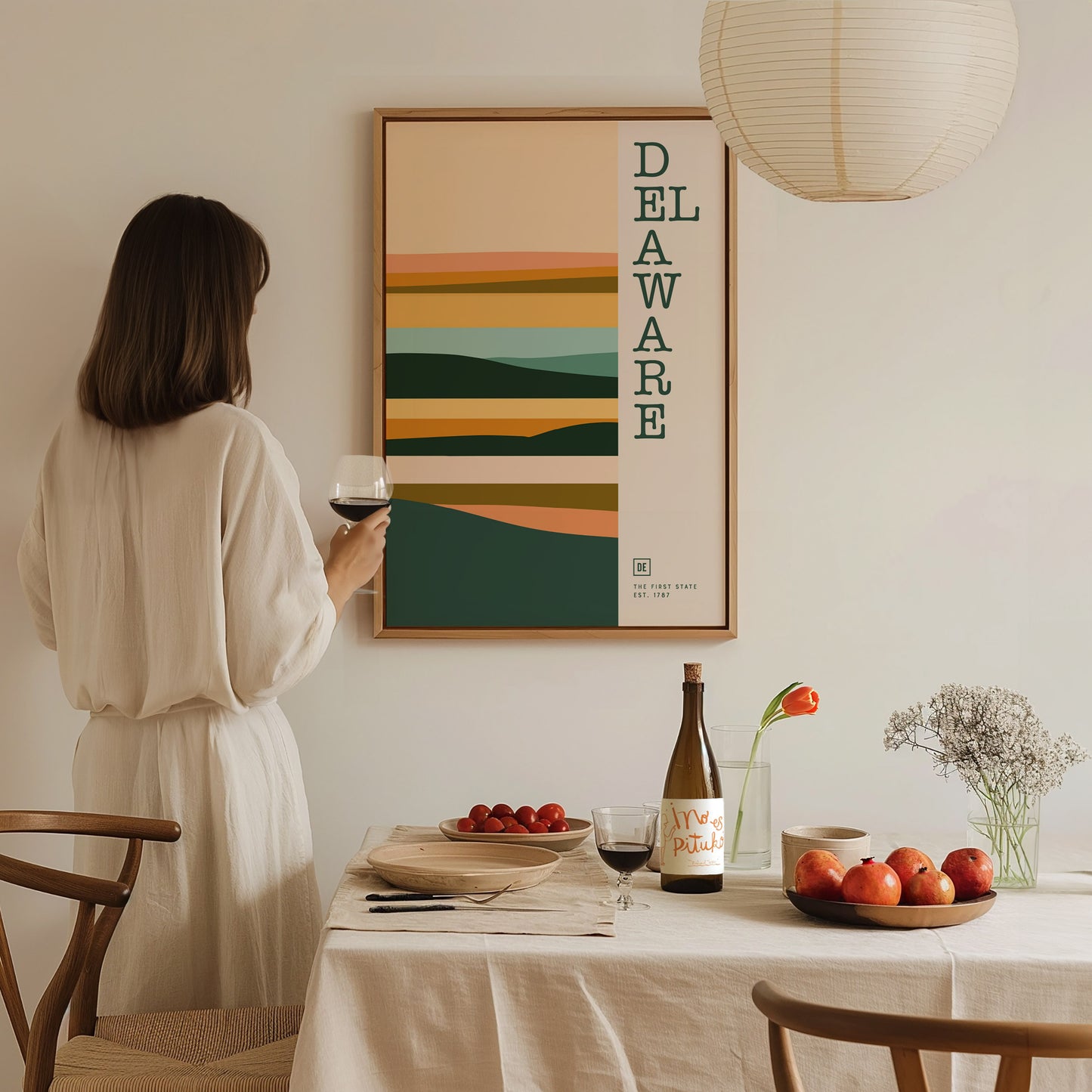 A woman in a white dress holds a glass of wine, admiring a Delaware-themed minimalist poster on the wall. A table with fruits, a wine bottle, and flowers is in.