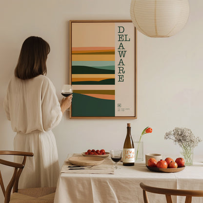 A woman in a white dress holds a glass of wine, admiring a Delaware-themed minimalist poster on the wall. A table with fruits, a wine bottle, and flowers is in.