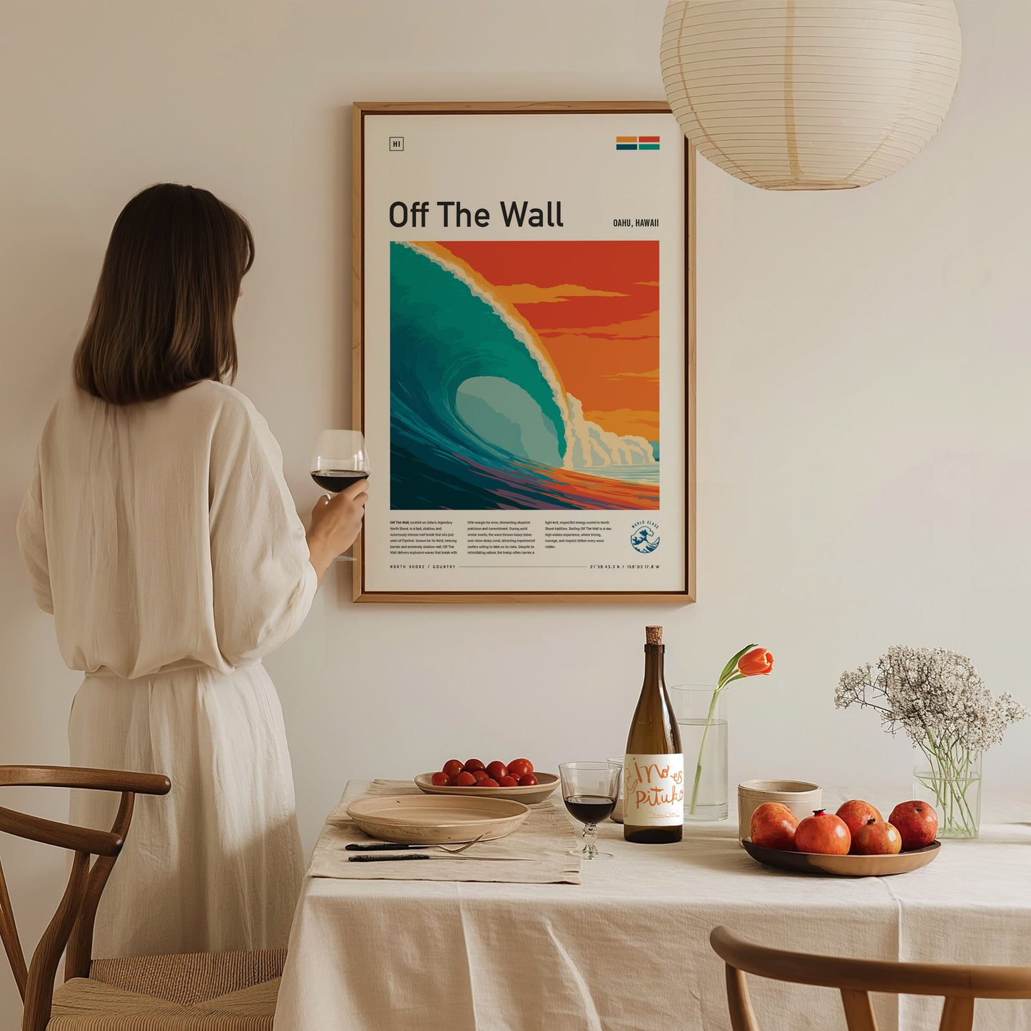 A woman in a light-colored, flowing dress holds a glass of wine while admiring a colorful poster titled Off The Wall, featuring a stylized wave against a sunset backdrop.