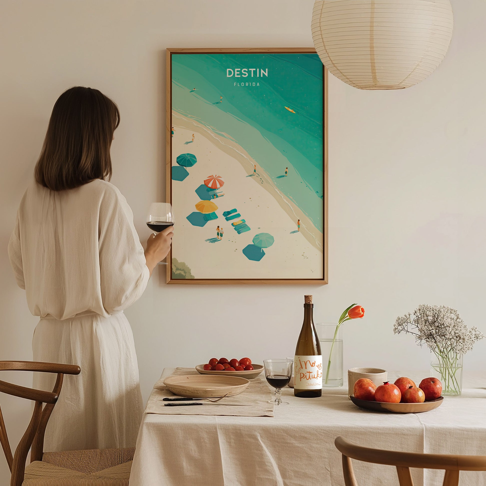 A woman in a white dress holds a glass of wine, admiring a Destin, Florida poster. The table is set with fruits, wine, and a flower vase.