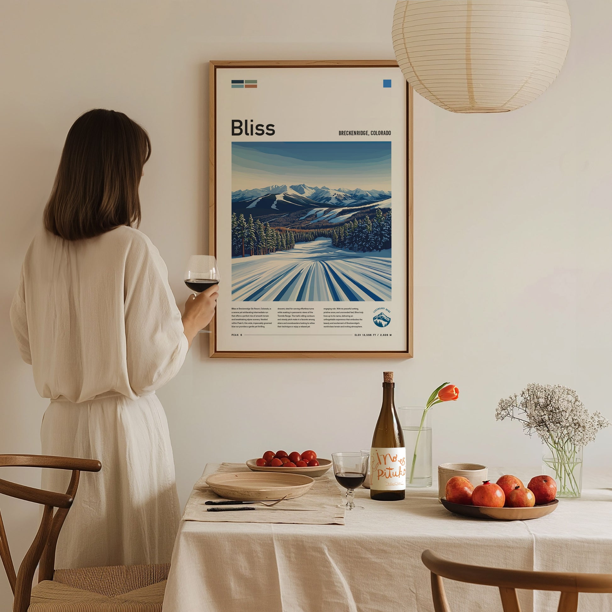 A woman in a white dress holds a glass of wine, admiring a framed Bliss ski run poster on the wall.