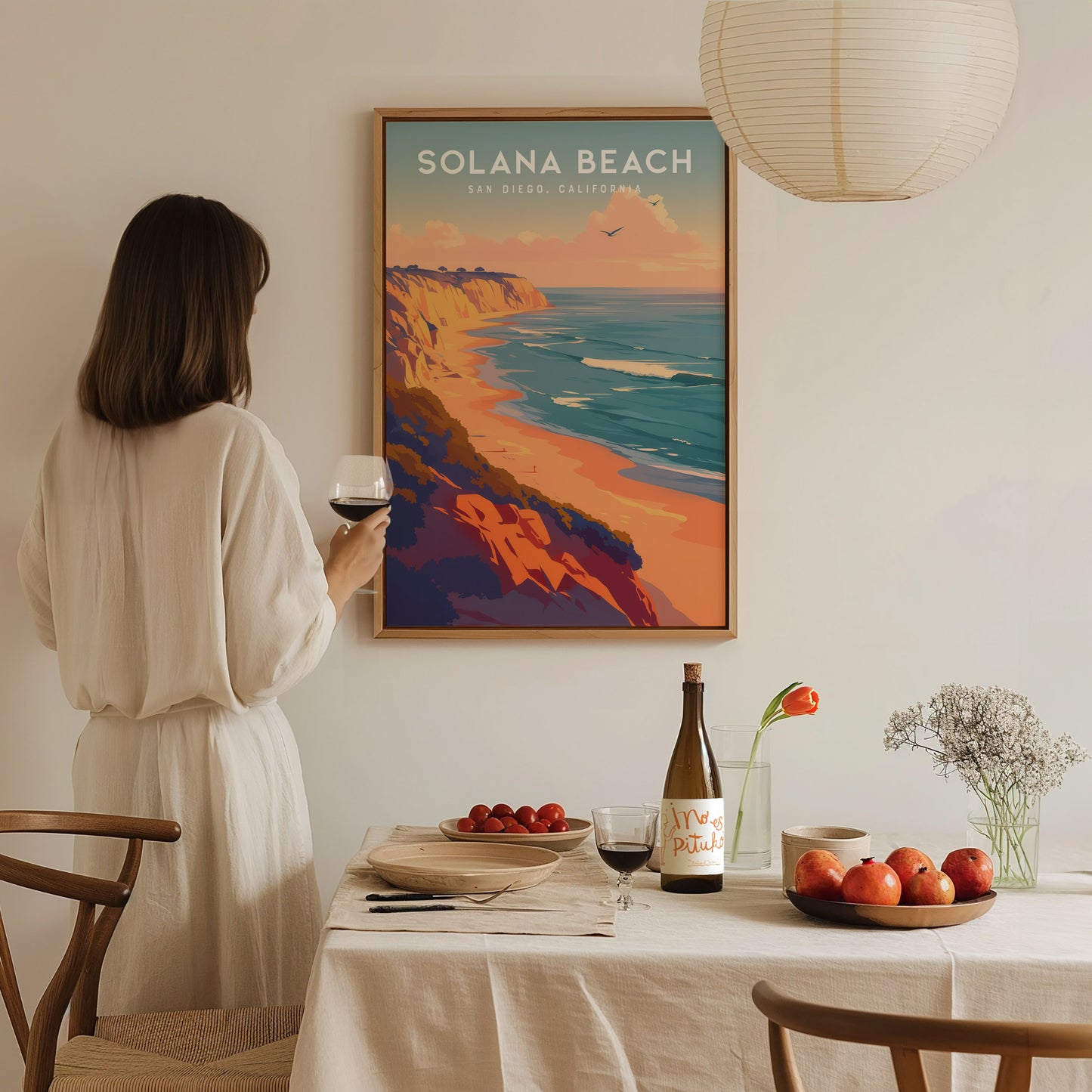 A woman in a beige dress holds a glass of wine, admiring a Solana Beach poster. The table is set with a bottle, fruit, and flowers, creating a cozy ambiance.