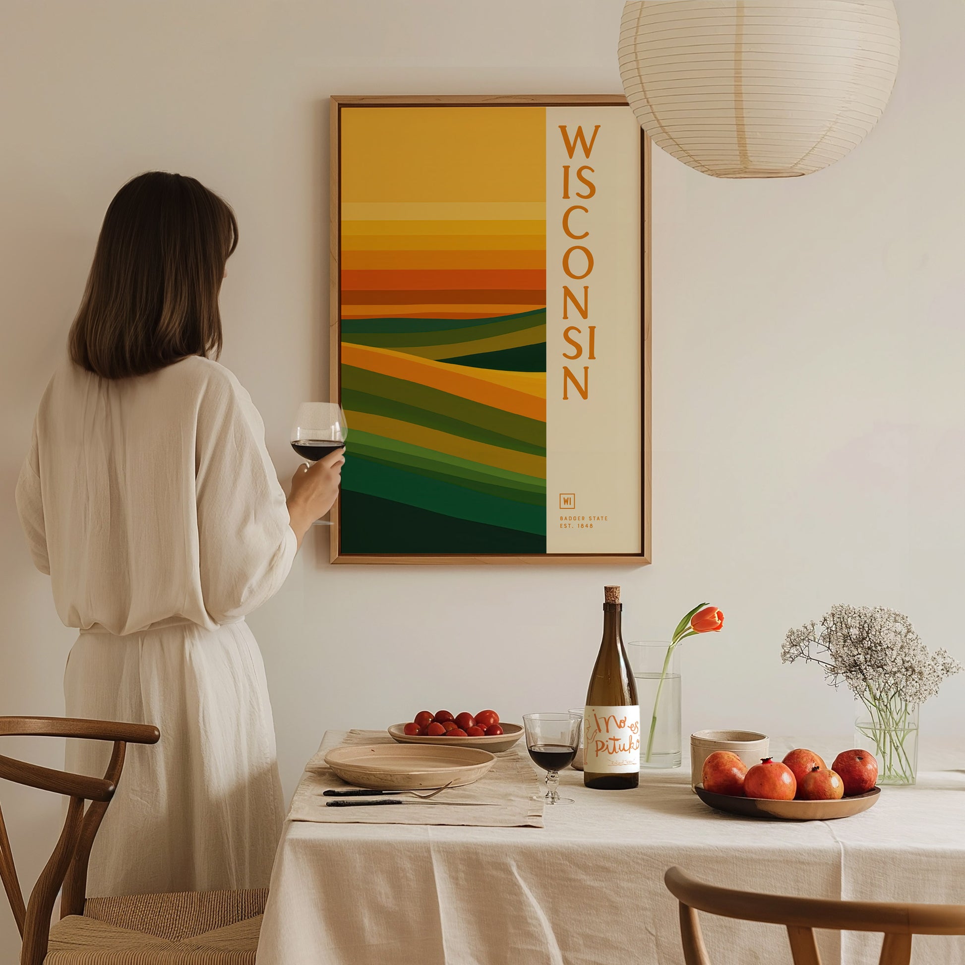 A woman holding a glass of wine admires a Wisconsin poster with abstract green and gold layers. The dining table is set with fruit, a bottle, and flowers.