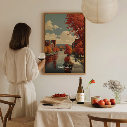 A woman in a white dress stands by a dining table, holding a wine glass, while gazing at a colorful poster of Bangor featuring autumn trees and a riverside scene.